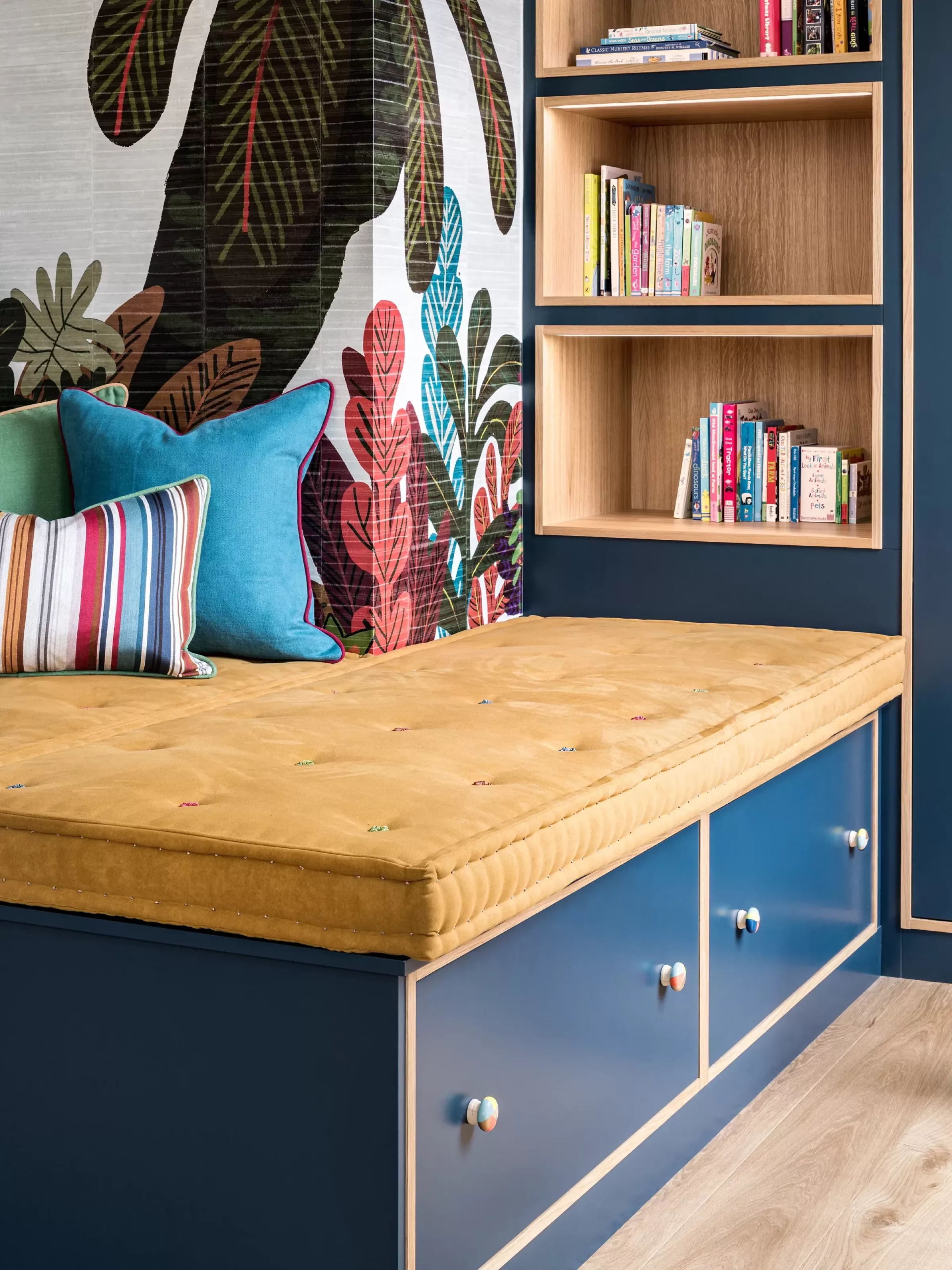 Built-in blue daybed with a mustard-yellow mattress and colourful striped and turquoise cushions set against tropical leaf-print wallpaper next to oak cubby shelves stocked with children's books