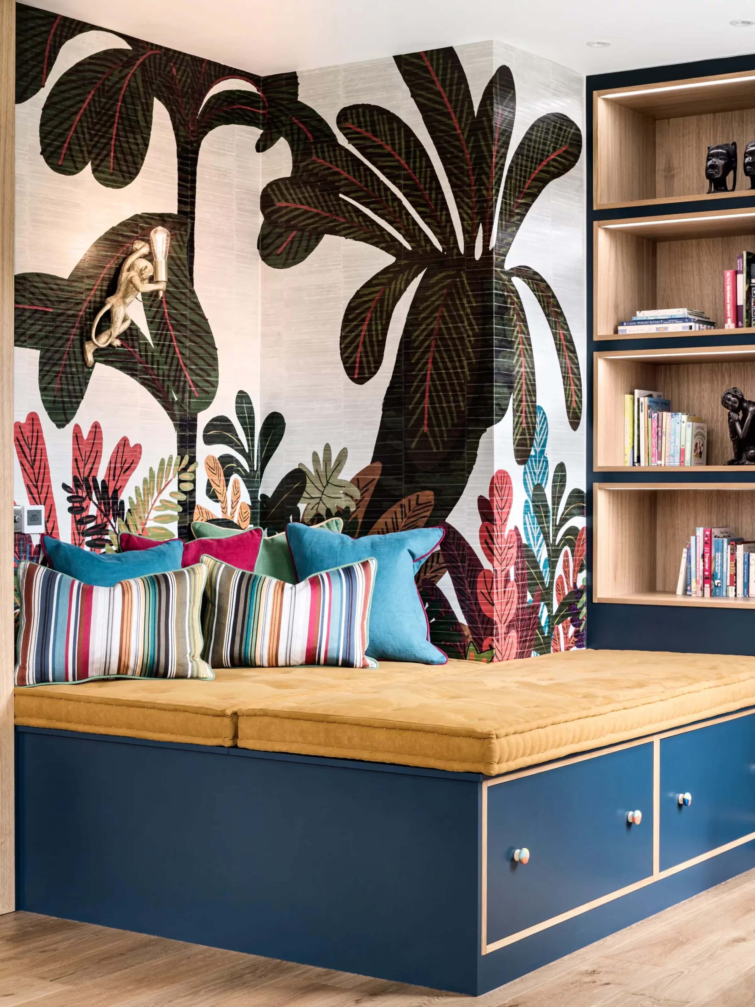 Built-in seating nook with a mustard cushion atop blue storage drawers, striped and jewel-toned throw pillows, a large tropical leaf mural on the corner walls, and adjacent wooden open shelving with books and small sculptures.