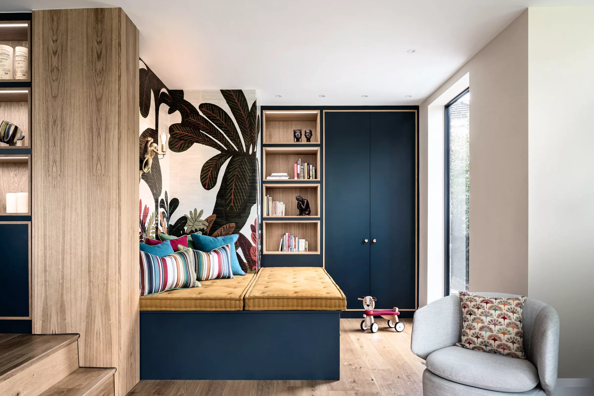 Built-in navy and oak reading nook with a mustard tufted bench topped with colourful striped cushions, open wooden shelves and cabinets beside a large botanical mural, a grey armchair with patterned cushion and a small wooden pull-along dog toy on the floor by a tall window.