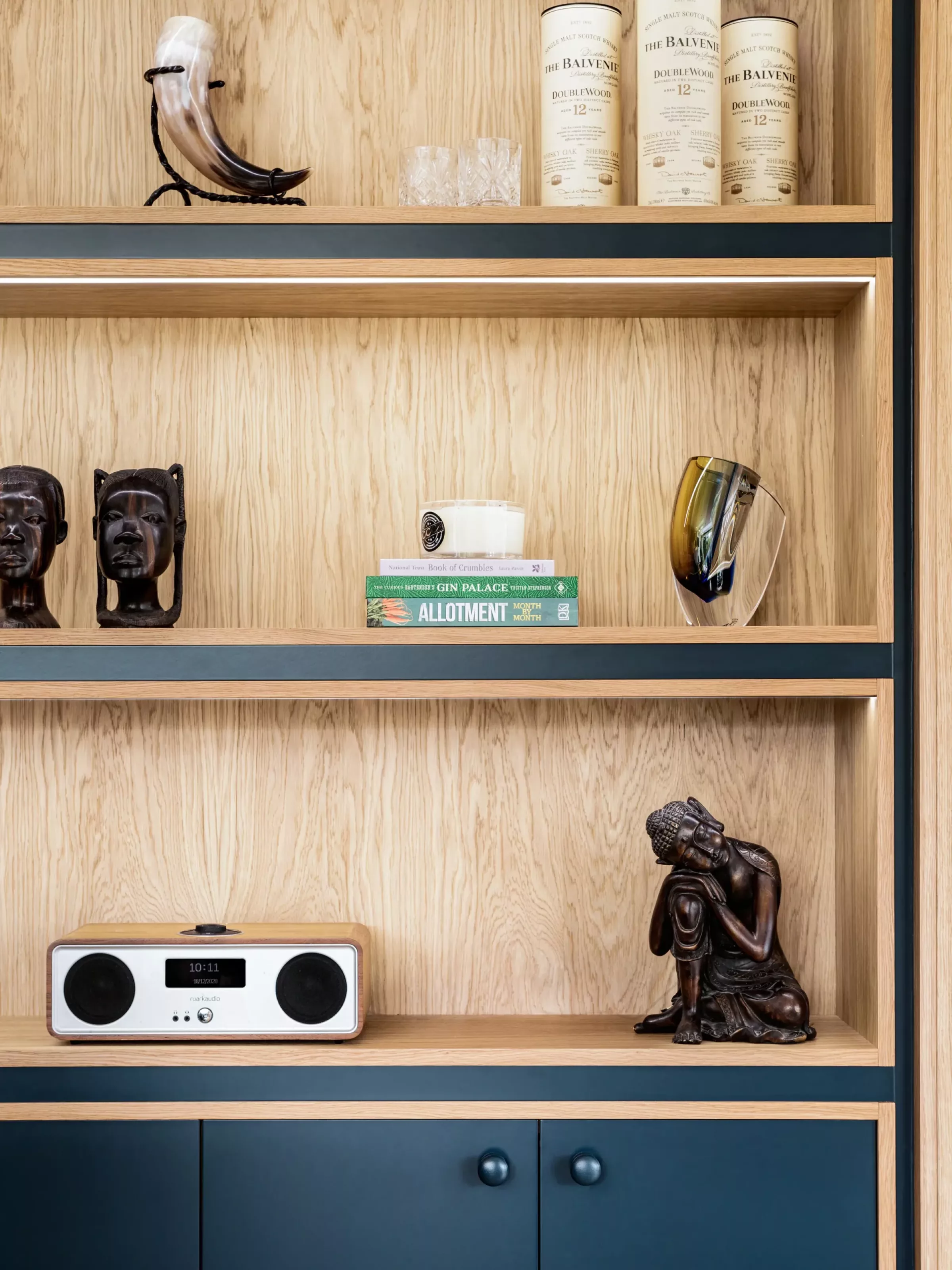 Light oak built-in shelving with navy-blue trim displaying decorative objects: top shelf with a drinking horn and whisky canisters, middle shelf with two carved wooden busts, a stack of green books topped by a candle and a tilted amber glass sculpture, and lower shelf with a compact radio and a seated wooden Buddha figure, with navy-blue cabinets beneath.