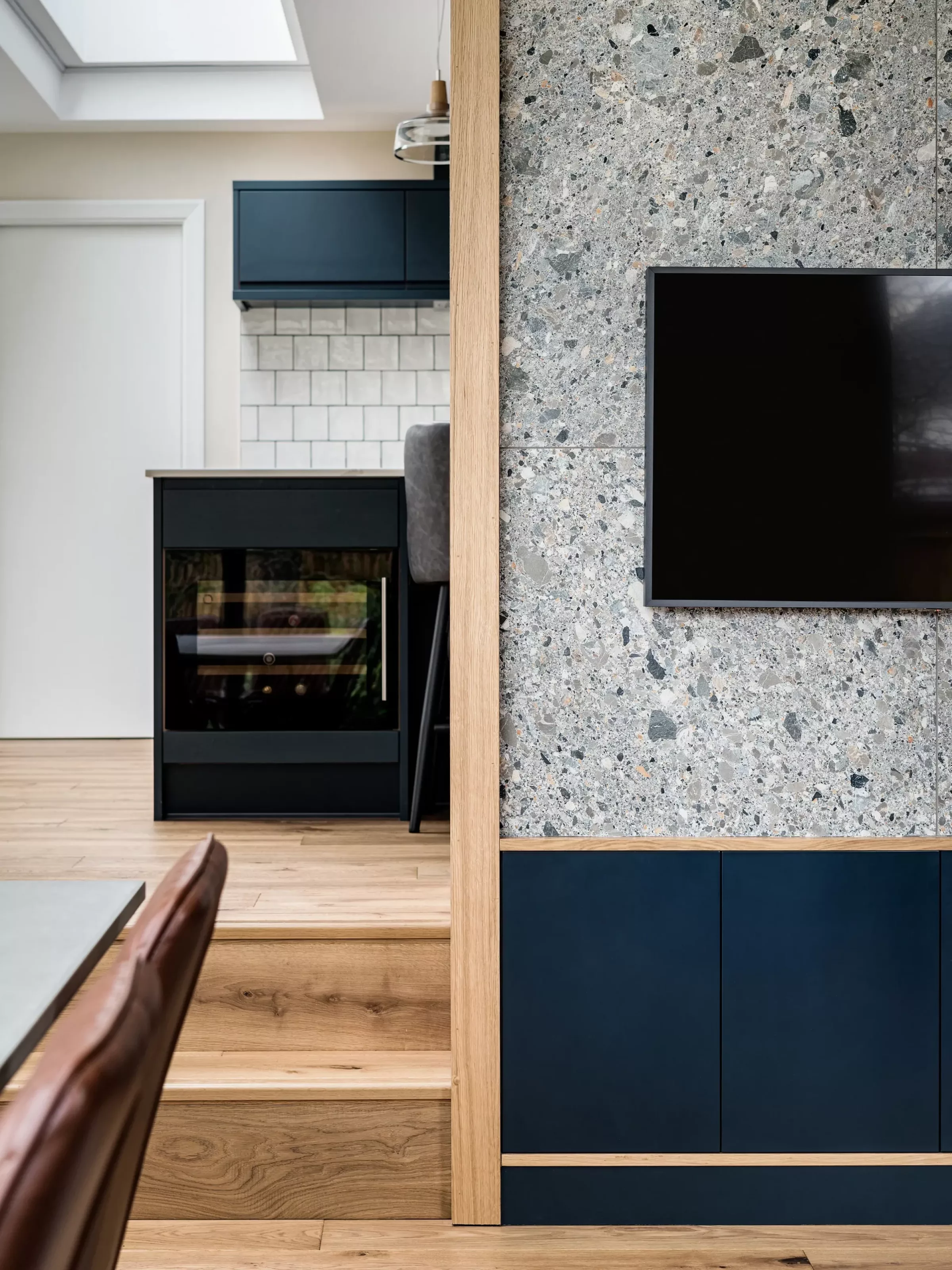 Split interior view showing a terrazzo-clad wall with oak trim and a wall-mounted black TV on the right, and navy kitchen cabinetry with a tiled backsplash, a black built-in appliance and oak steps leading to a dining area beneath a skylight on the left.