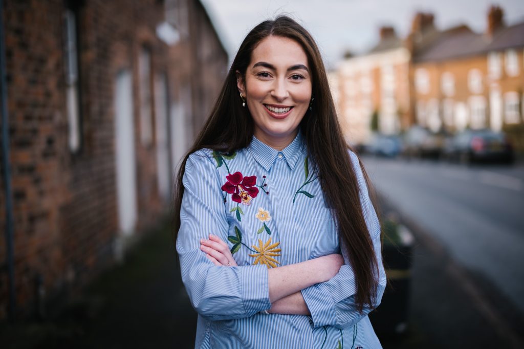 Smiling woman in a floral embroidered shirt standing on a residential street.