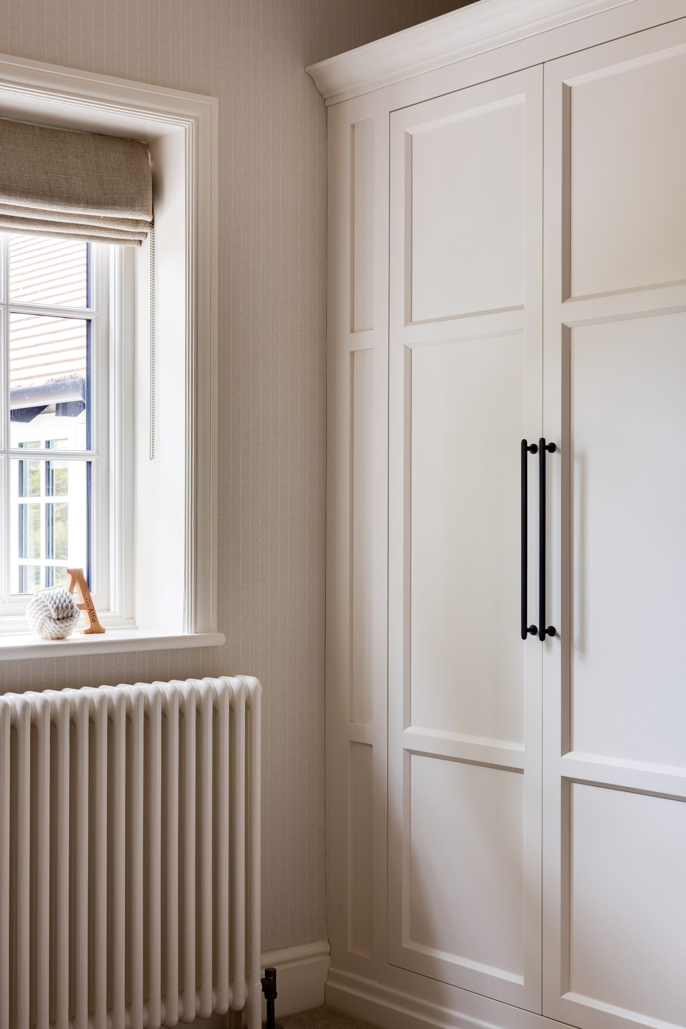 A built-in wardrobe with panelled cream doors and black handles, next to a window with a roman blind and a traditional white radiator.