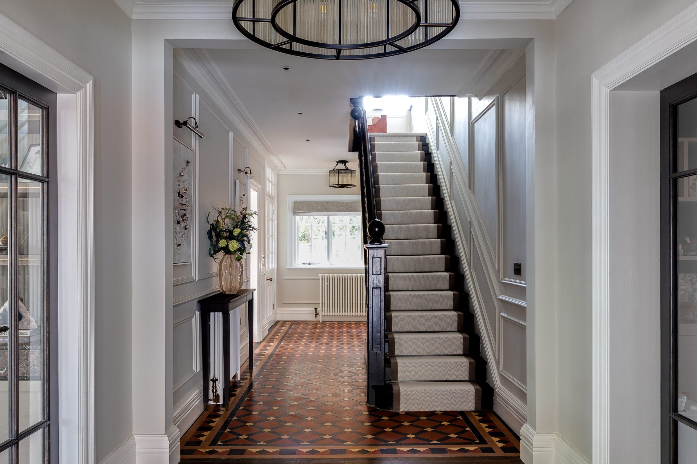 Grand hallway with geometric floor tiles and dark timber staircase with a central carpet runner, framed by soft wall panelling.