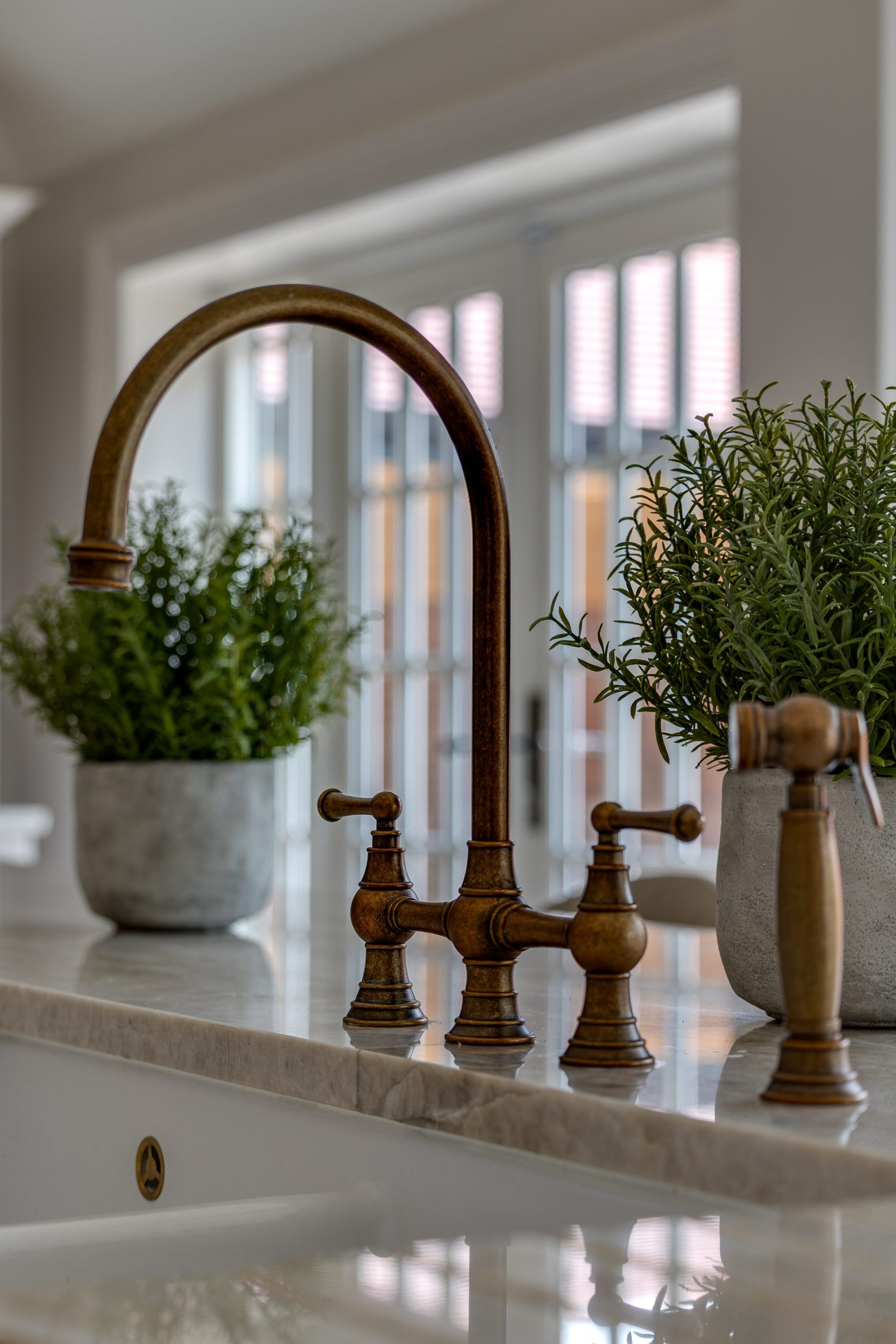 A brass kitchen tap on a marble countertop, framed by two potted green plants in stone pots.