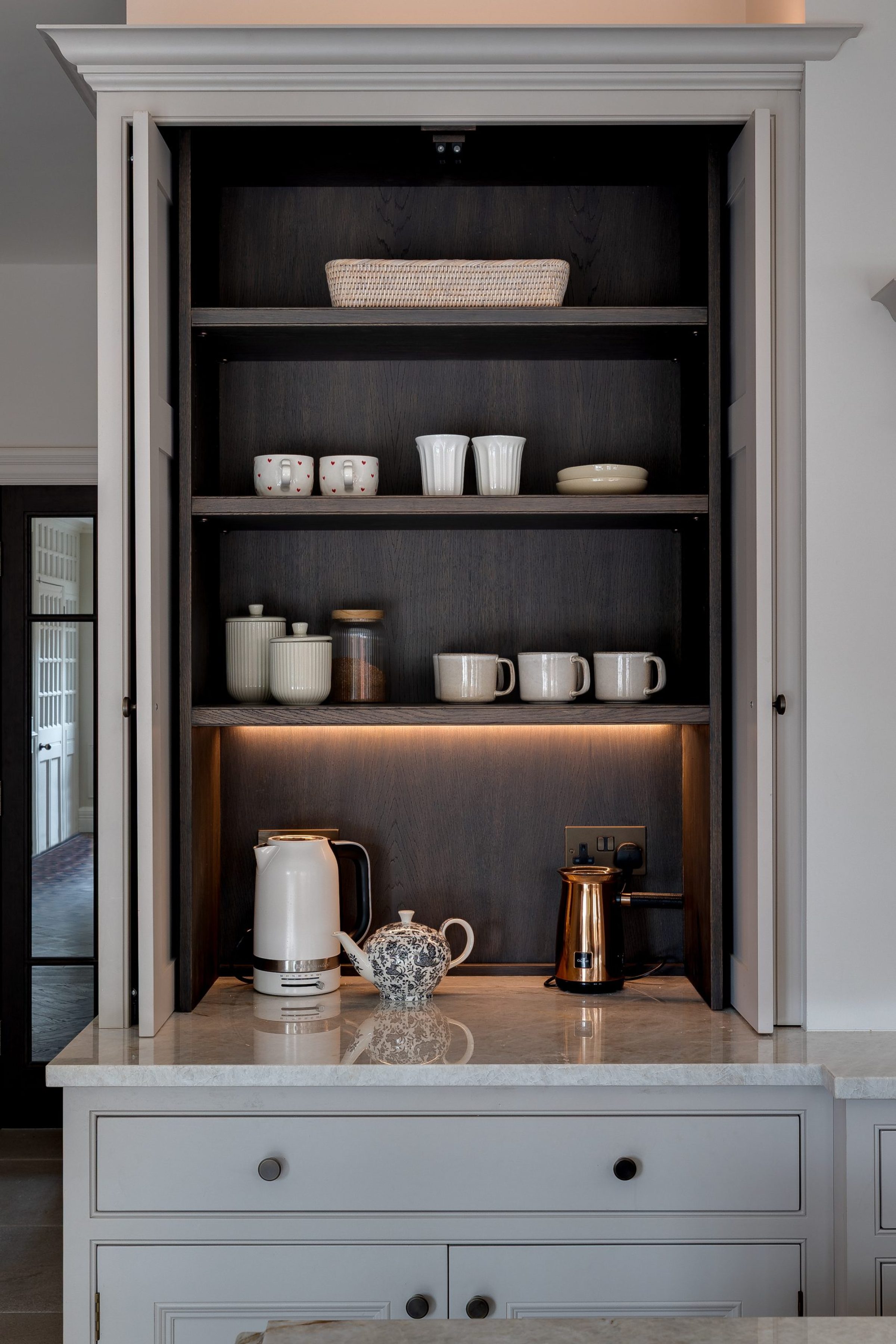 Open built-in kitchen larder with dark wood shelves, mugs, jars and a kettle on a marble countertop.