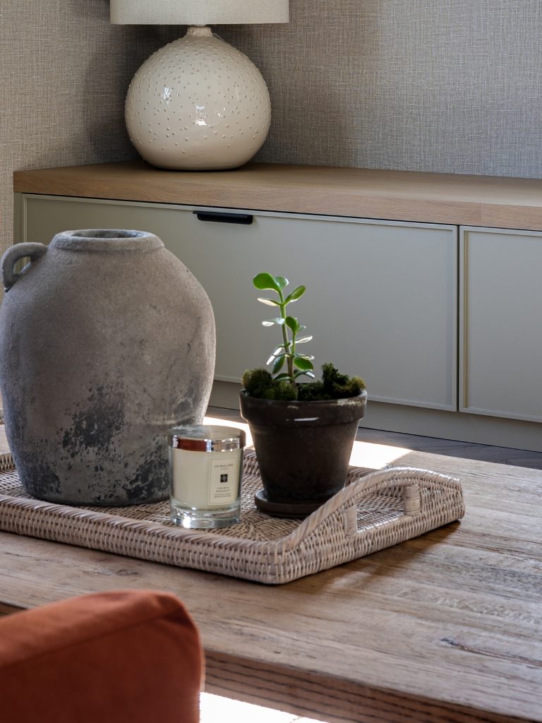 Wicker tray with jug, candle, and plant on wooden coffee table in a neutral-toned living room.