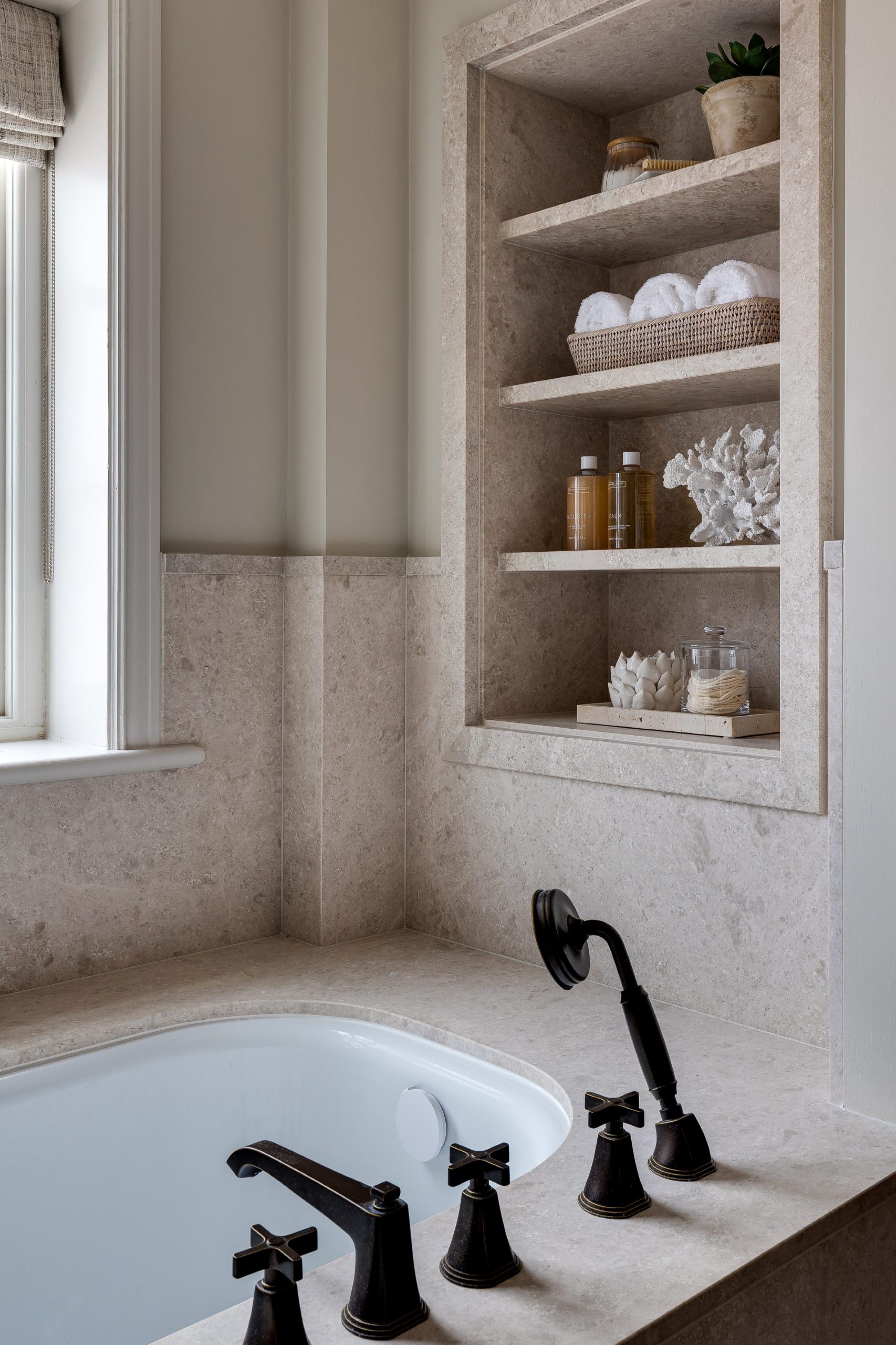 Built-in stone shelves styled with towels, coral, and accessories above a bath with black crosshead taps and a handheld shower.