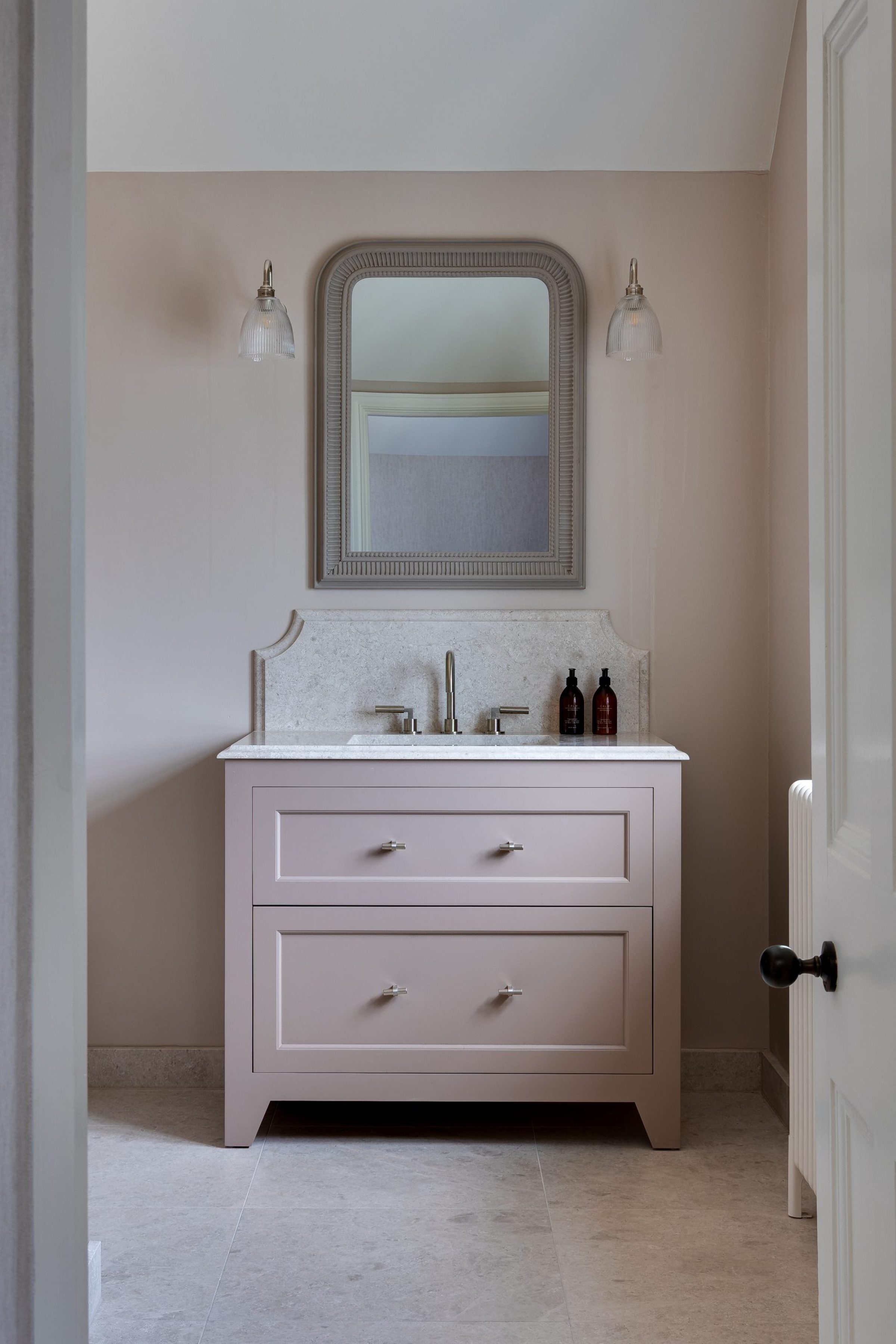 Soft pink bathroom vanity with stone countertop, wall mirror, and glass wall lights on either side.