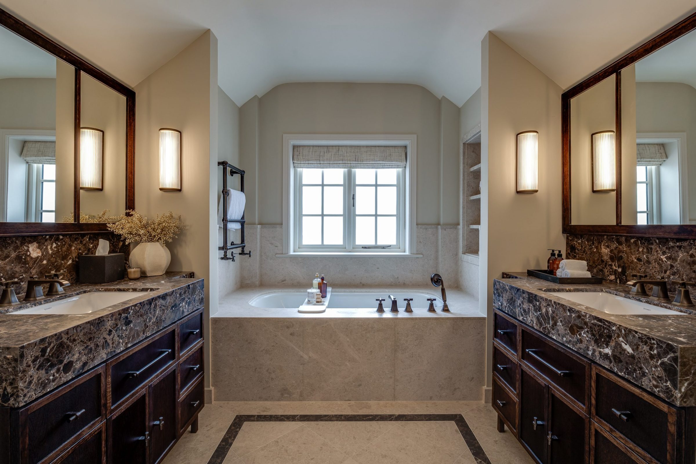 A luxury master bathroom with a central bathtub framed by twin vanities, featuring dark marble countertops, integrated shelving, and soft wall lighting.