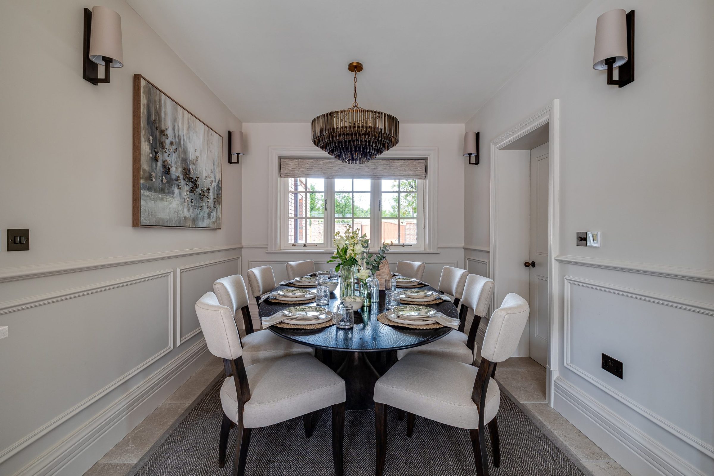 A dining room with a large round dark wood table set for eight, surrounded by upholstered cream chairs, with a modern chandelier overhead, wall panelling, and a large window bringing in natural light.