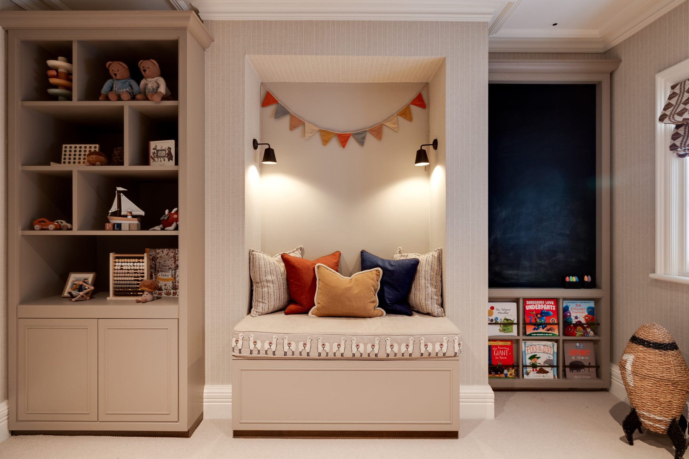 A cosy children’s reading nook with built-in shelving, playful cushions, wall lighting, and a blackboard surrounded by books and toys.