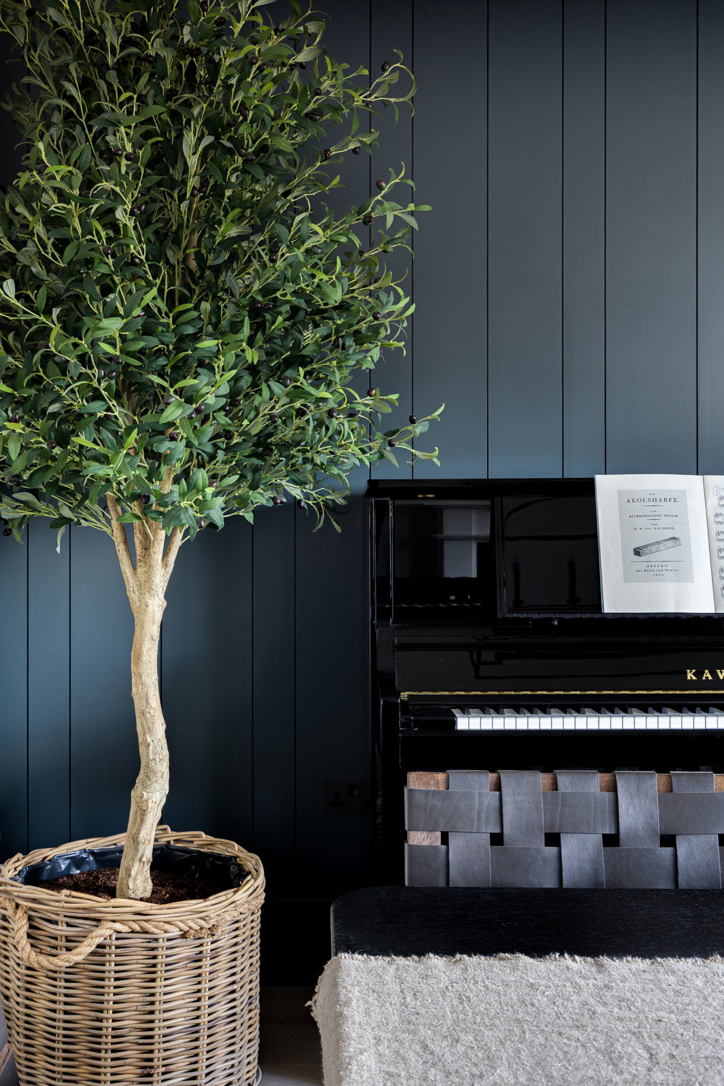 Decorative corner featuring a black upright piano with sheet music and a potted indoor tree in a woven basket against dark paneling.