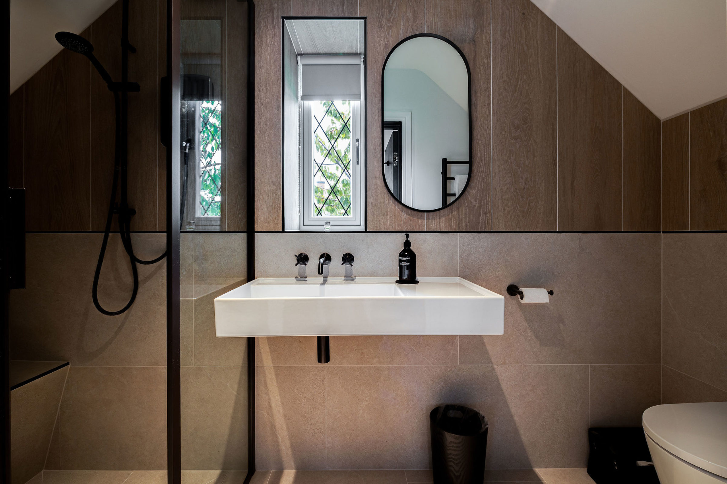 Contemporary attic bathroom with wood paneling, wall-mounted sink, black fixtures, and glass shower enclosure.