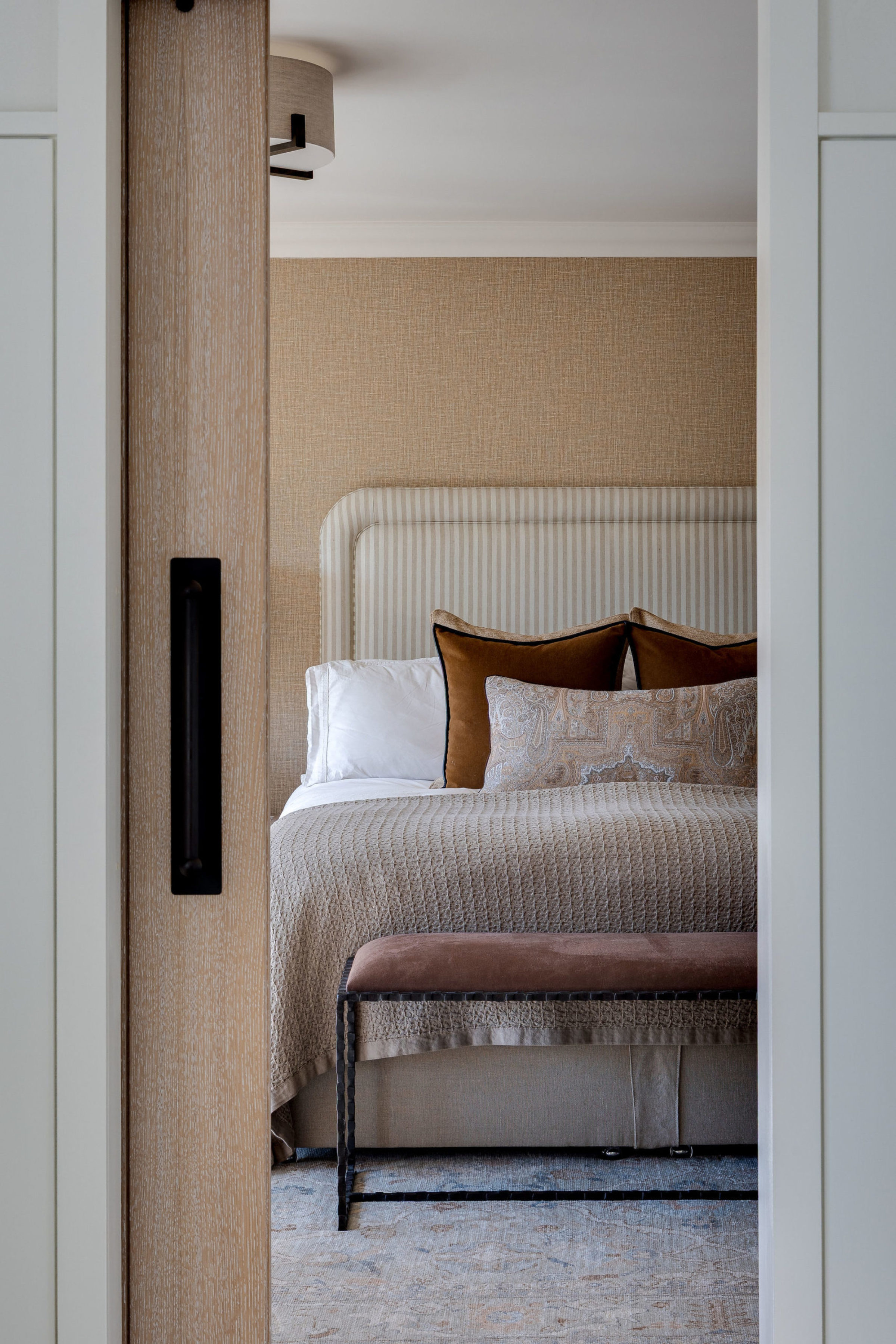 View through sliding door into a serene bedroom with neutral tones, striped headboard, and layered cushions.