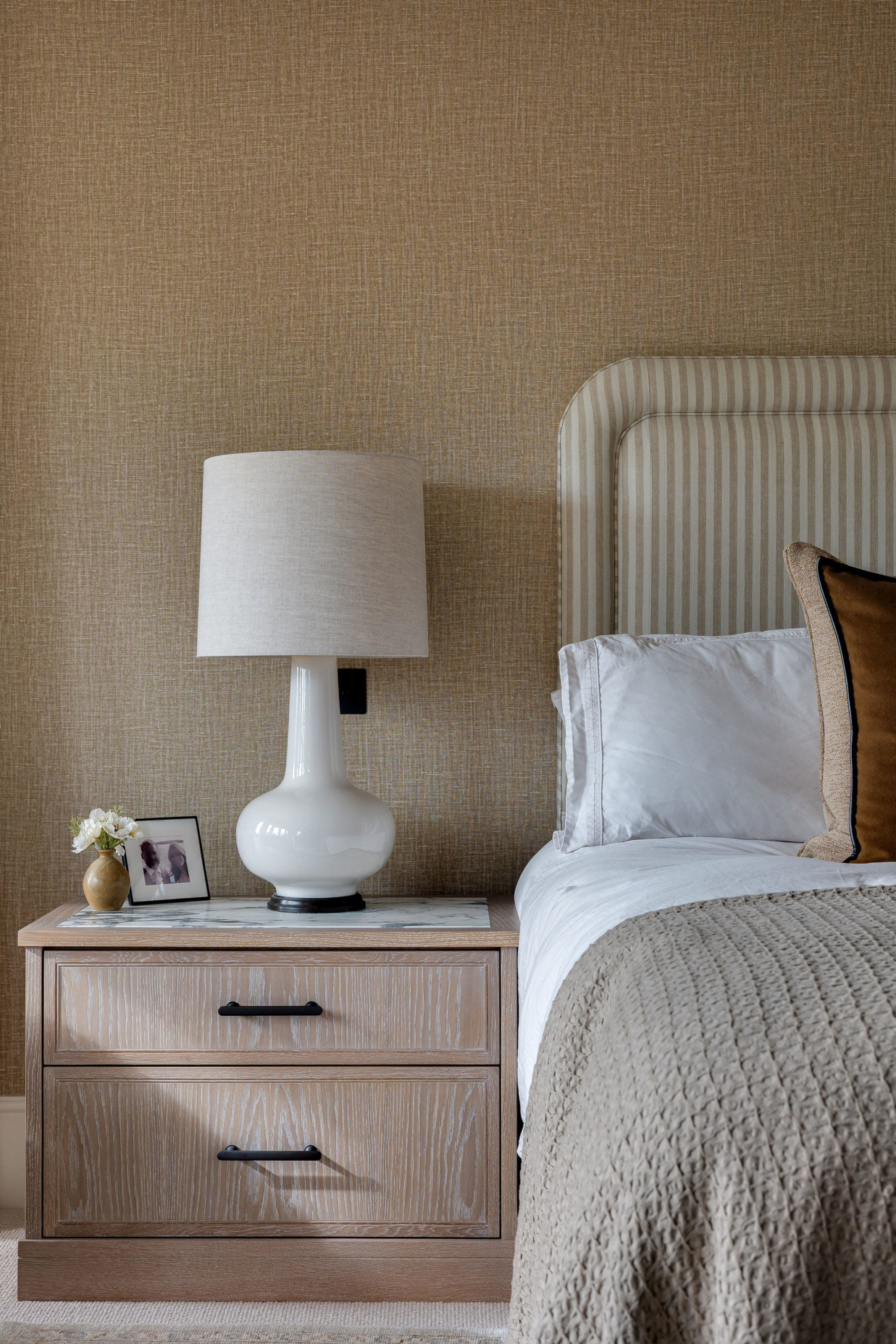 Bedside table with white ceramic lamp, framed photo, and neutral upholstered headboard.