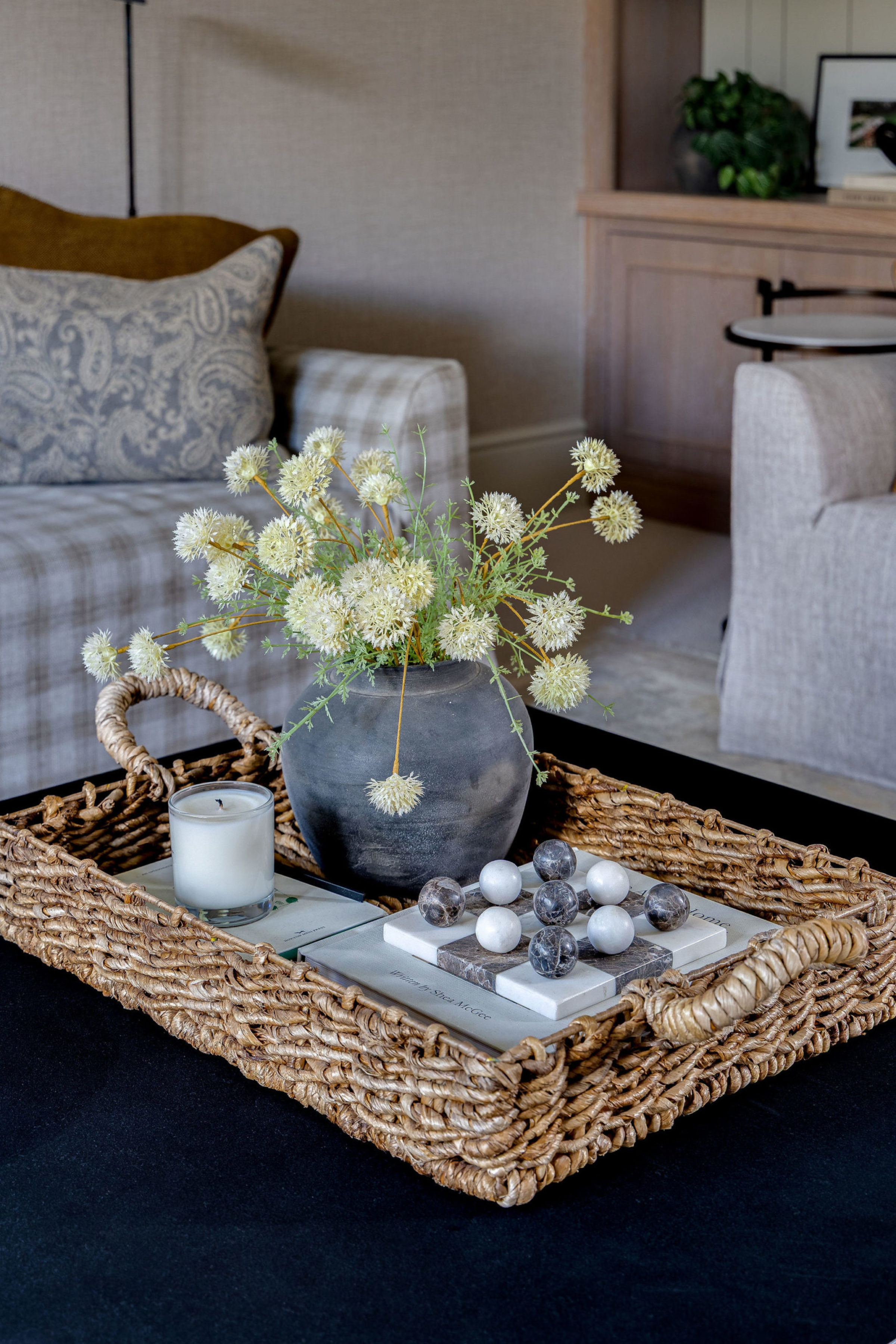 Close-up of woven tray on coffee table with ceramic vase of white flowers, candle, and decorative marble spheres.