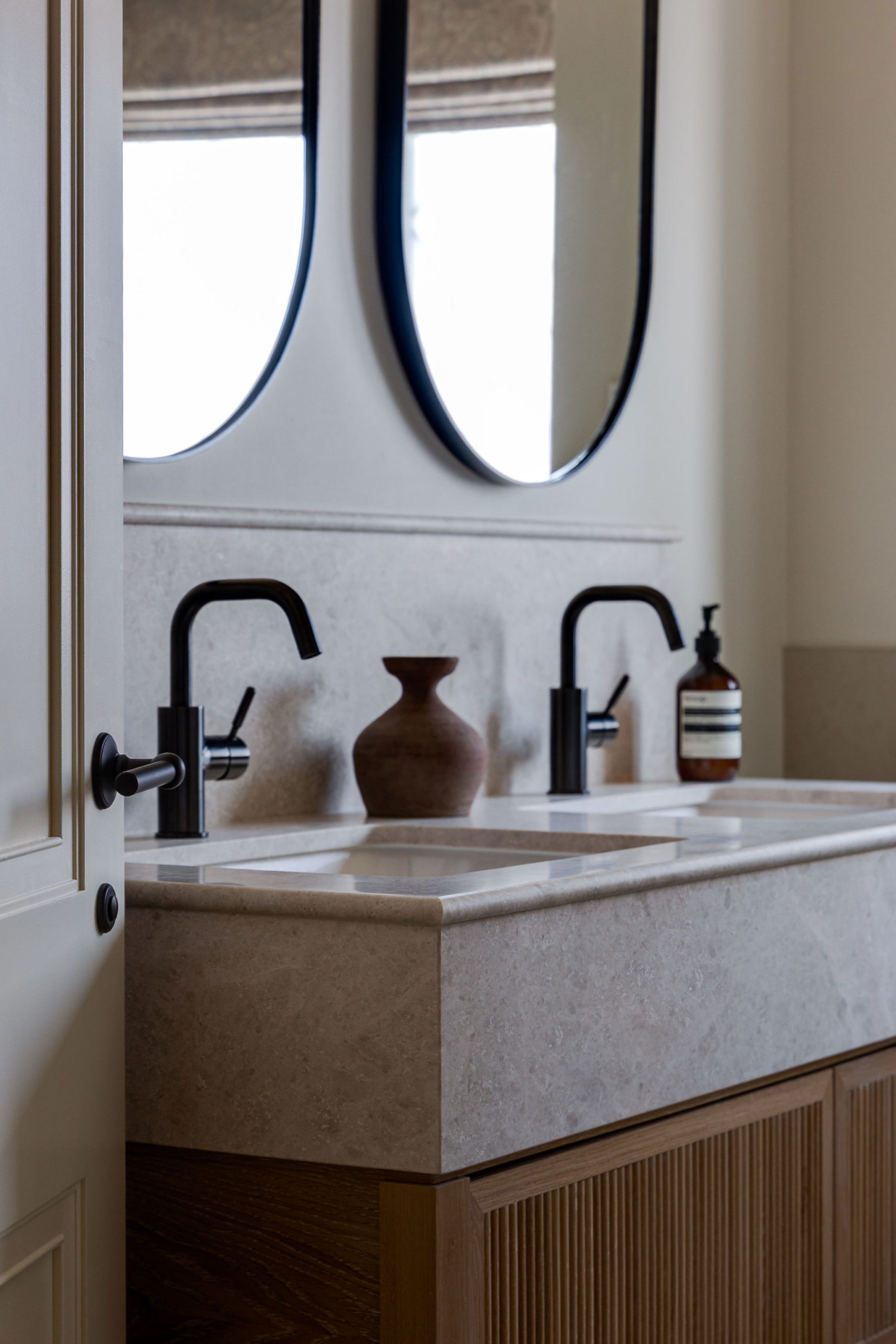 Elegant guest bathroom vanity with twin sinks, black faucets, and oval mirrors above stone countertop.