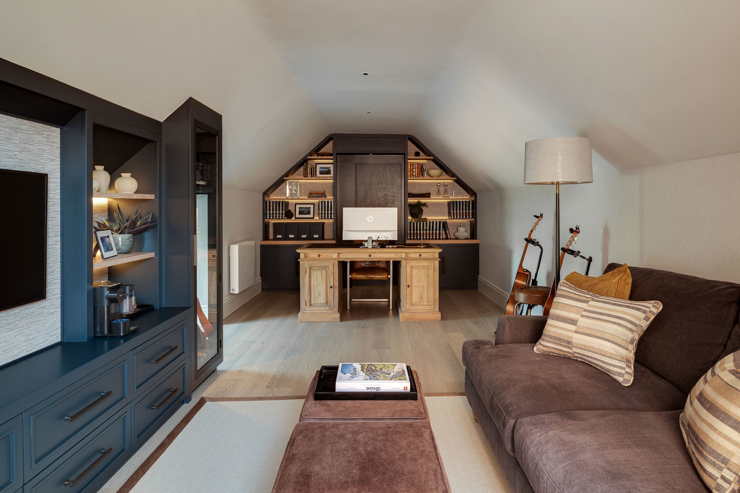 Attic home office with built-in shelving, wooden desk, and cozy seating area with guitars.
