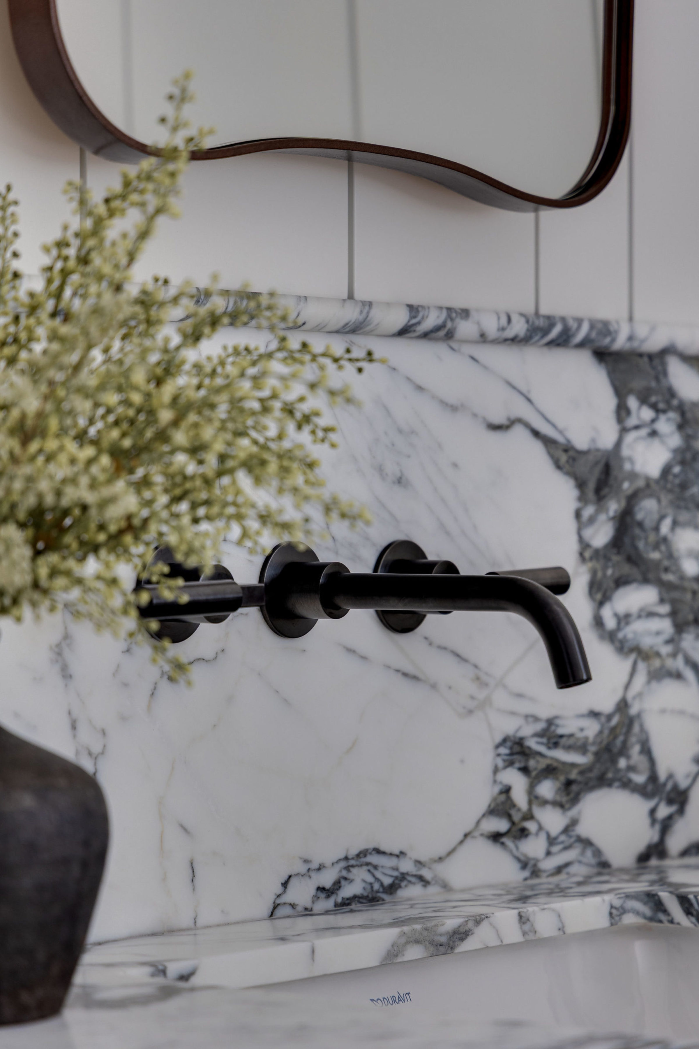 Detail of black wall-mounted faucet over white marble sink with decorative vase and greenery.