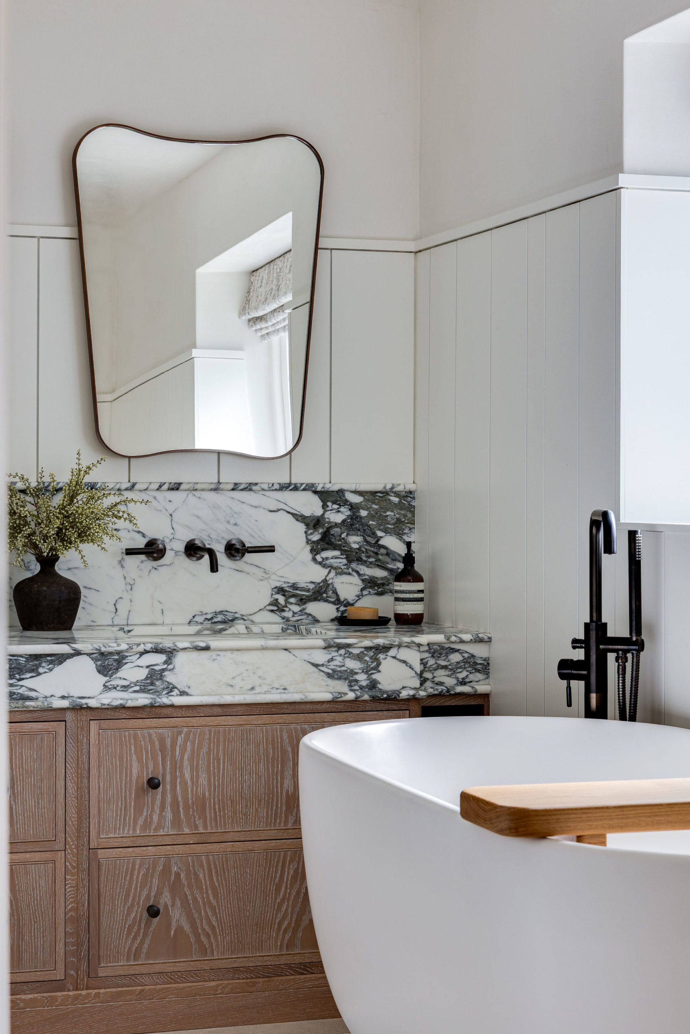 Close view of freestanding bathtub beside marble vanity with organic mirror and black fixtures.