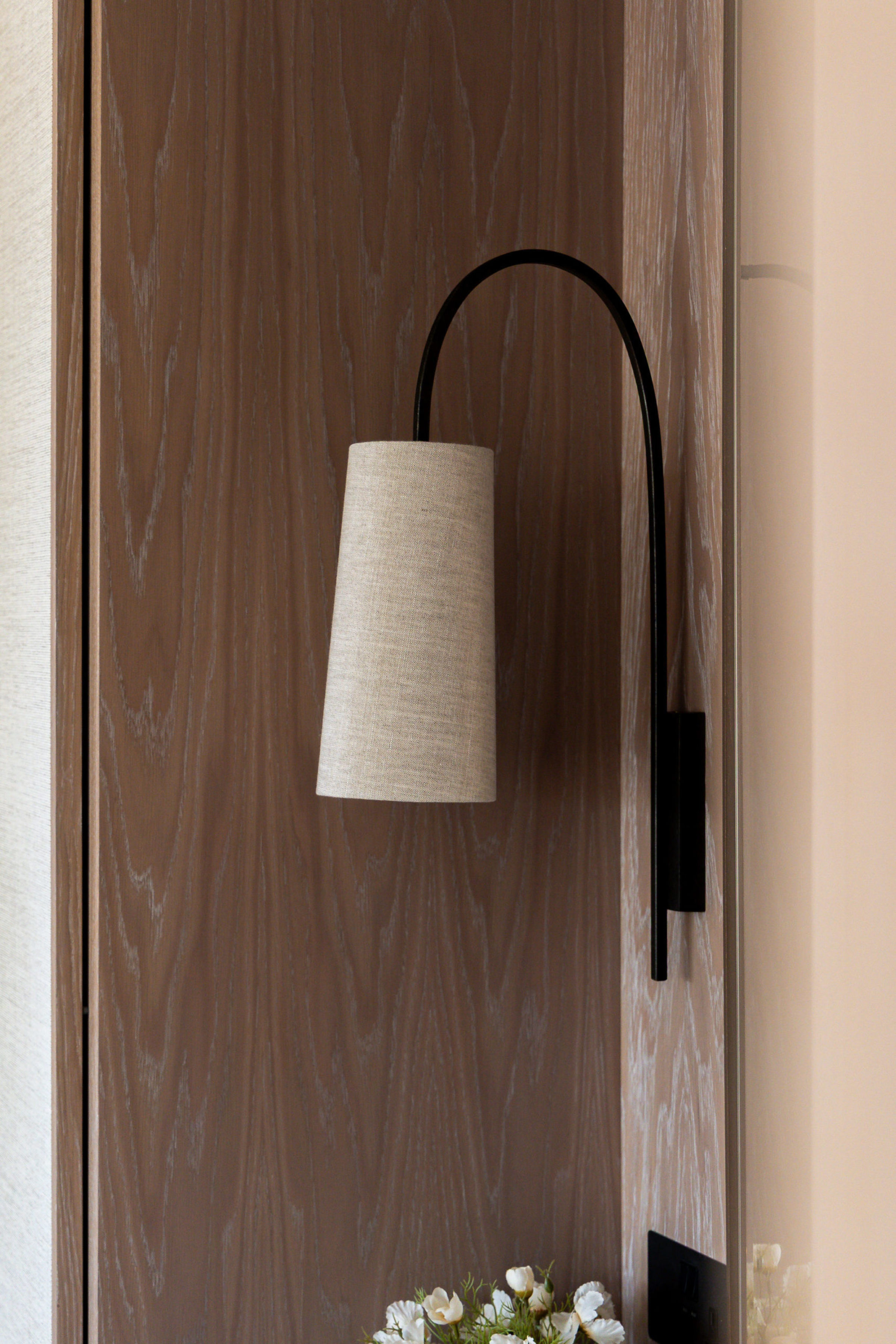 Close-up of modern black wall sconce with fabric shade mounted on light oak paneling above a floral arrangement.