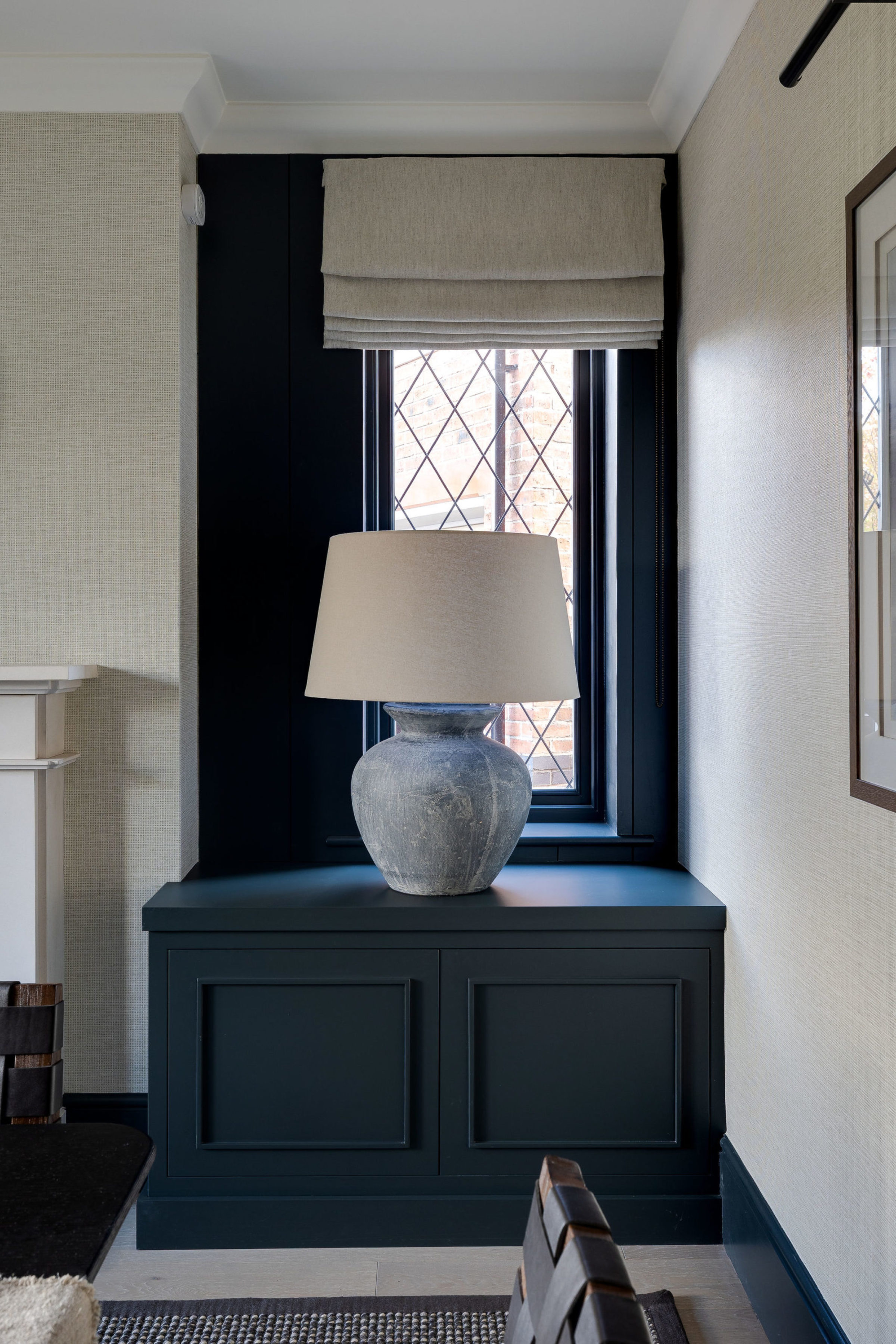 Cozy window nook with navy cabinetry, Roman shade, and textured ceramic table lamp.