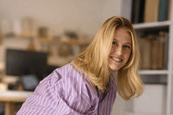 A smiling young woman with shoulder-length blonde hair, wearing a purple and white striped shirt, is leaning forward while working in a bright, modern office space filled with shelves and a computer in the background.