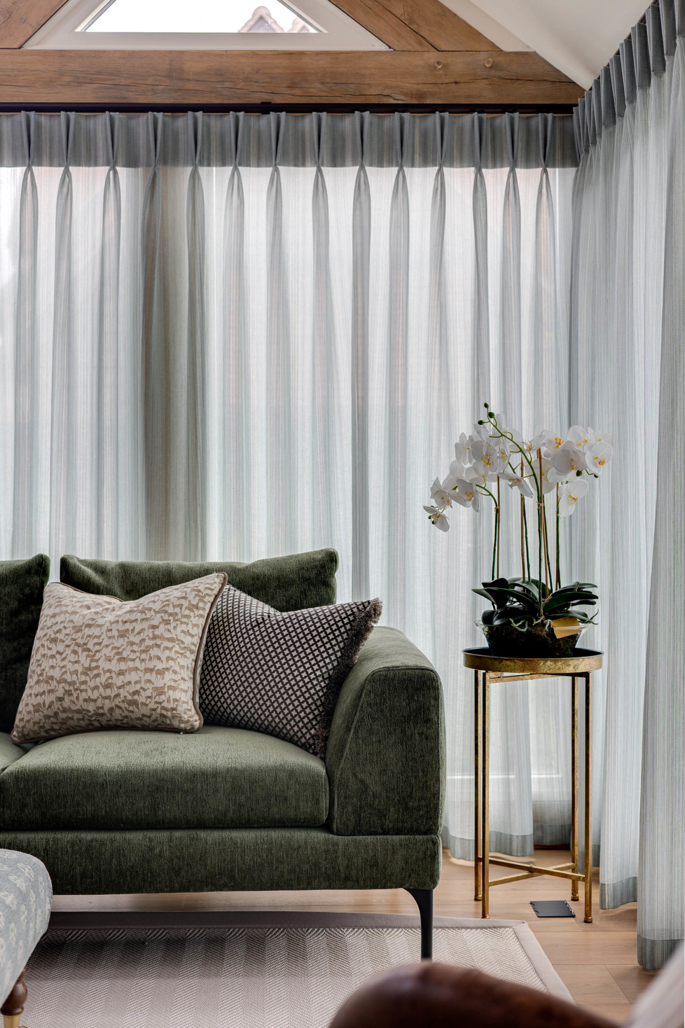 Green velvet sofa with patterned cushions beside a tall gold side table holding a white orchid, set in front of pale grey sheer curtains beneath a wooden beam and triangular window.