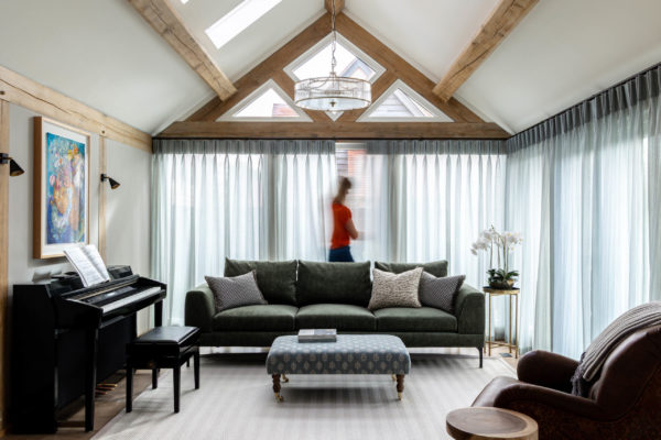 Vaulted living room framed by exposed timber beams and triangular clerestory windows, sheer floor-to-ceiling curtains backlighting a dark green sofa with patterned cushions and a blue patterned ottoman; a black piano with sheet music sits to the left, a drum-style chandelier hangs overhead, a small side table holds a white orchid, and a blurred figure walks behind the curtains.
