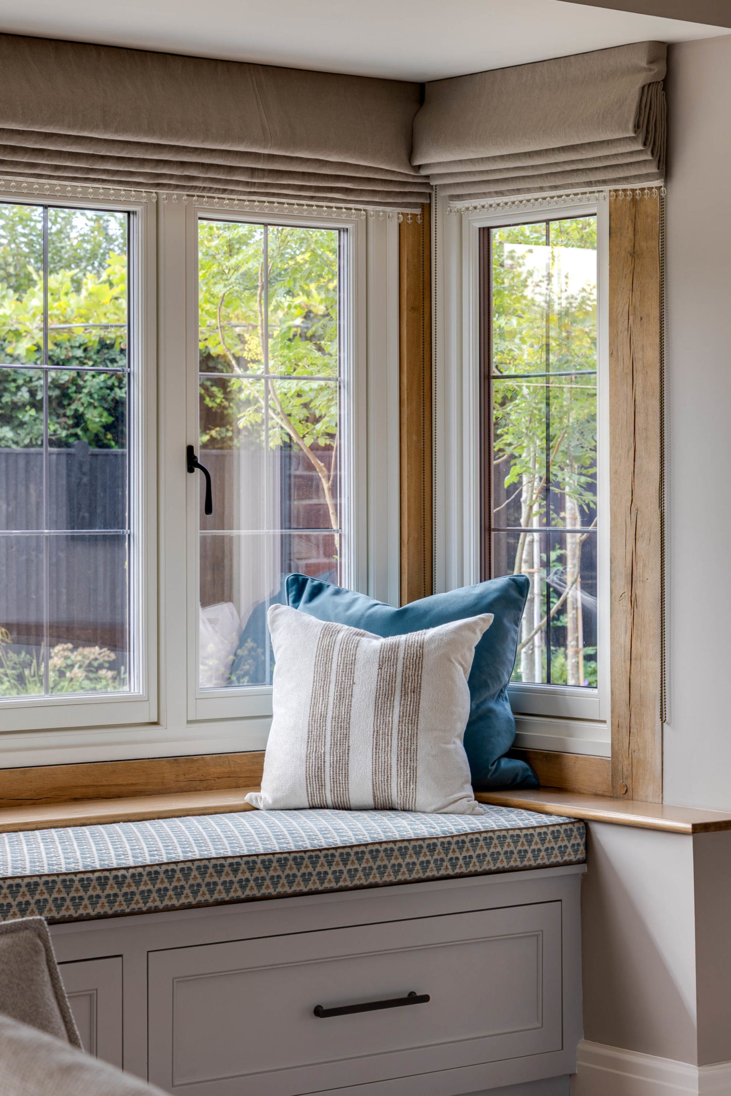 Cosy window seat in a bay with a patterned bench cushion, a cream striped throw pillow and a teal cushion, framed by white-framed windows and folded beige Roman blinds trimmed with small crystal beads, revealing a leafy garden outside.