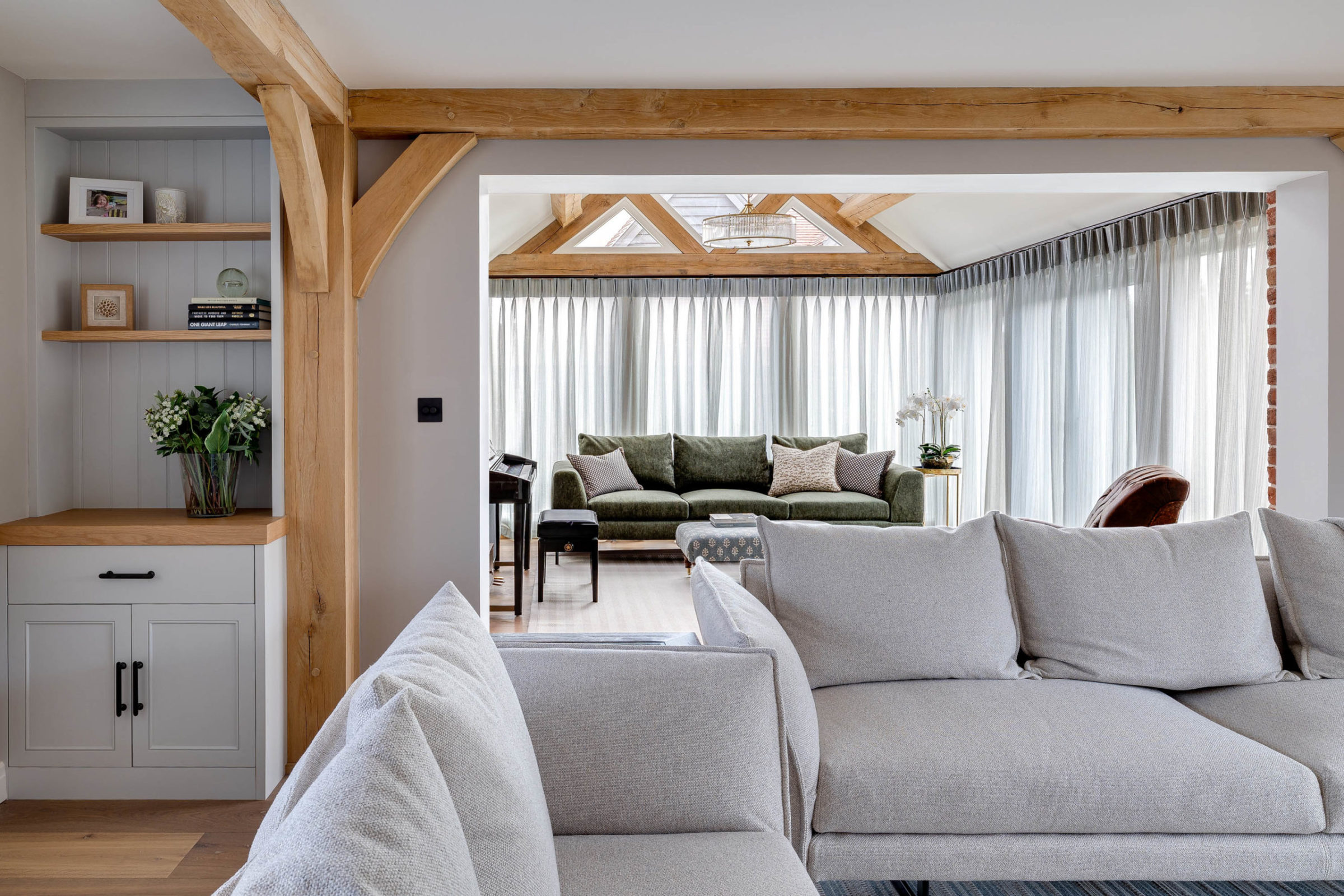 Open-plan living area with a light‑grey upholstered sectional in the foreground, oak beams and built‑in shelves to the left, and a sunlit adjoining room beyond containing a green velvet sofa, sheer floor‑to‑ceiling curtains, exposed wooden trusses and a round chandelier.