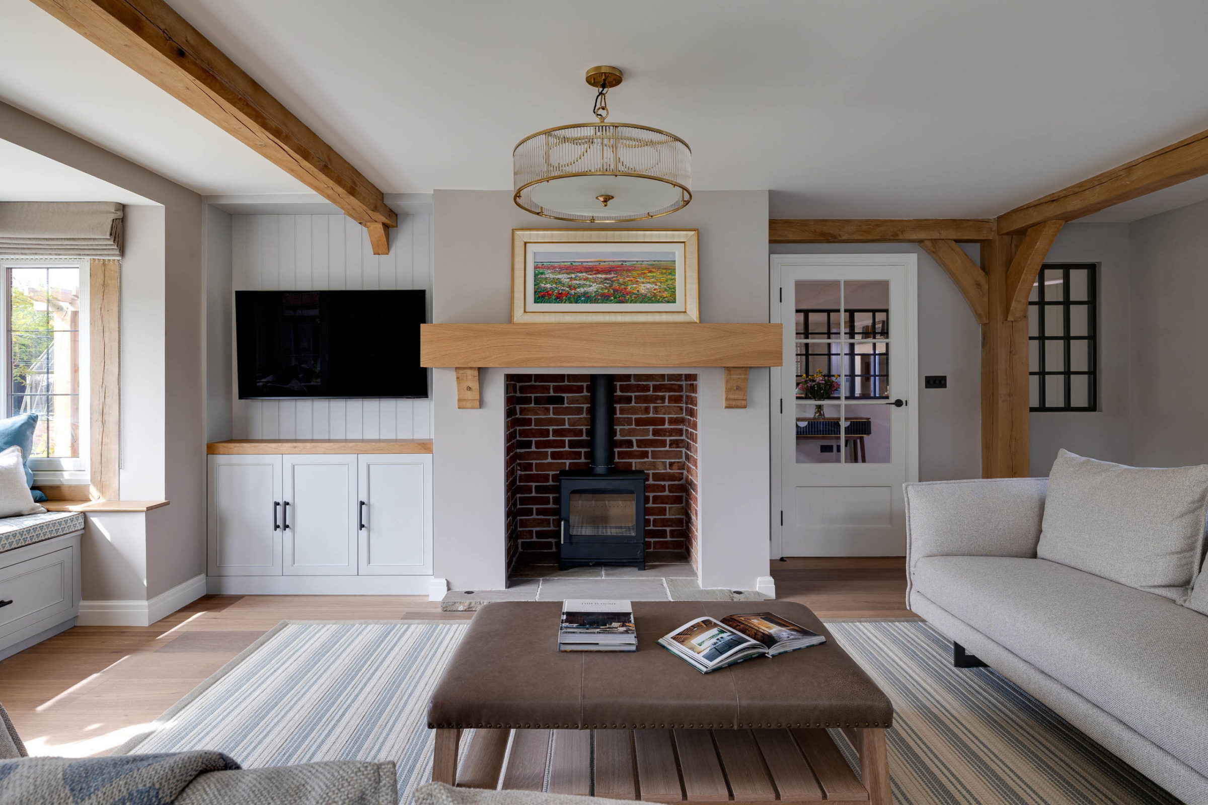 Cozy modern living room with exposed oak ceiling beams and a central brick fireplace topped by a wooden mantel and framed landscape painting, flanked by a wall-mounted TV and white built-in cabinets, with a glass‑paned door to the right, a brown ottoman with magazines on a striped rug and light grey sofas.