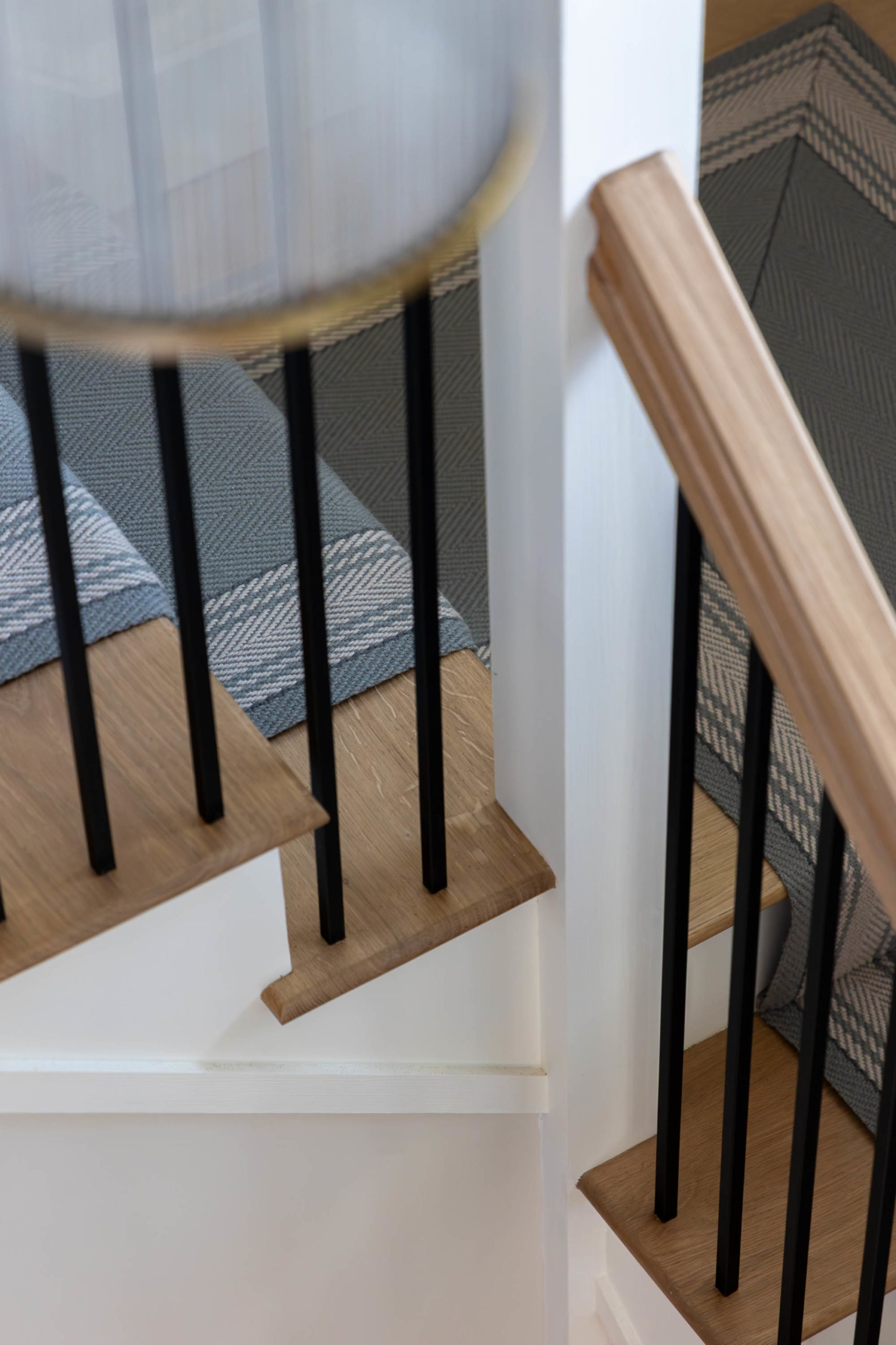 Close-up of a wooden staircase corner showing oak treads, a light wood handrail, vertical black metal balusters, a white newel post and a grey patterned stair runner.