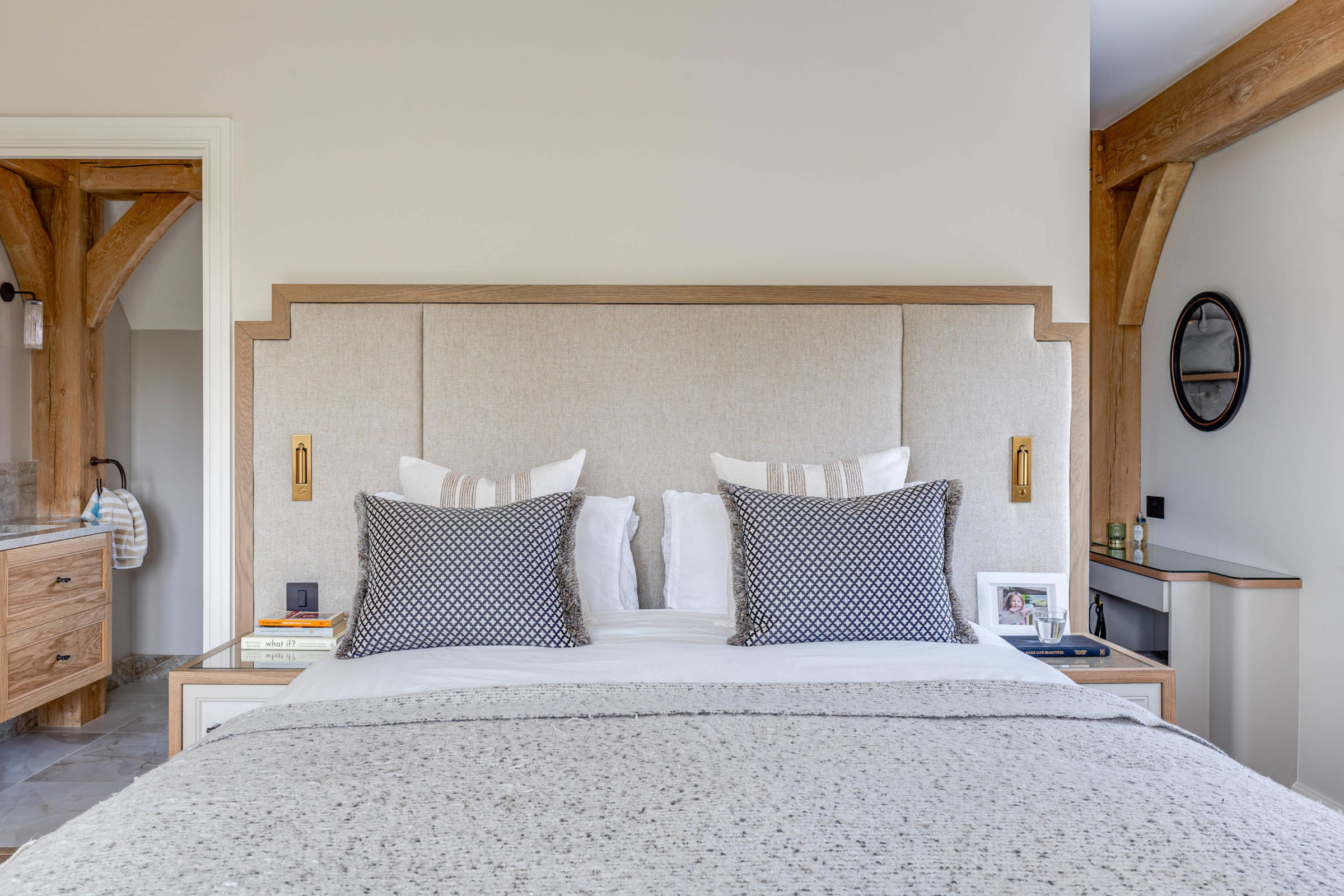 Neutral-toned bedroom with a large upholstered headboard framed in wood, two patterned square cushions and white pillows on the bed, exposed wooden beams, bedside tables holding books, a framed photo and a glass of water, and a small round mirror on the right wall.