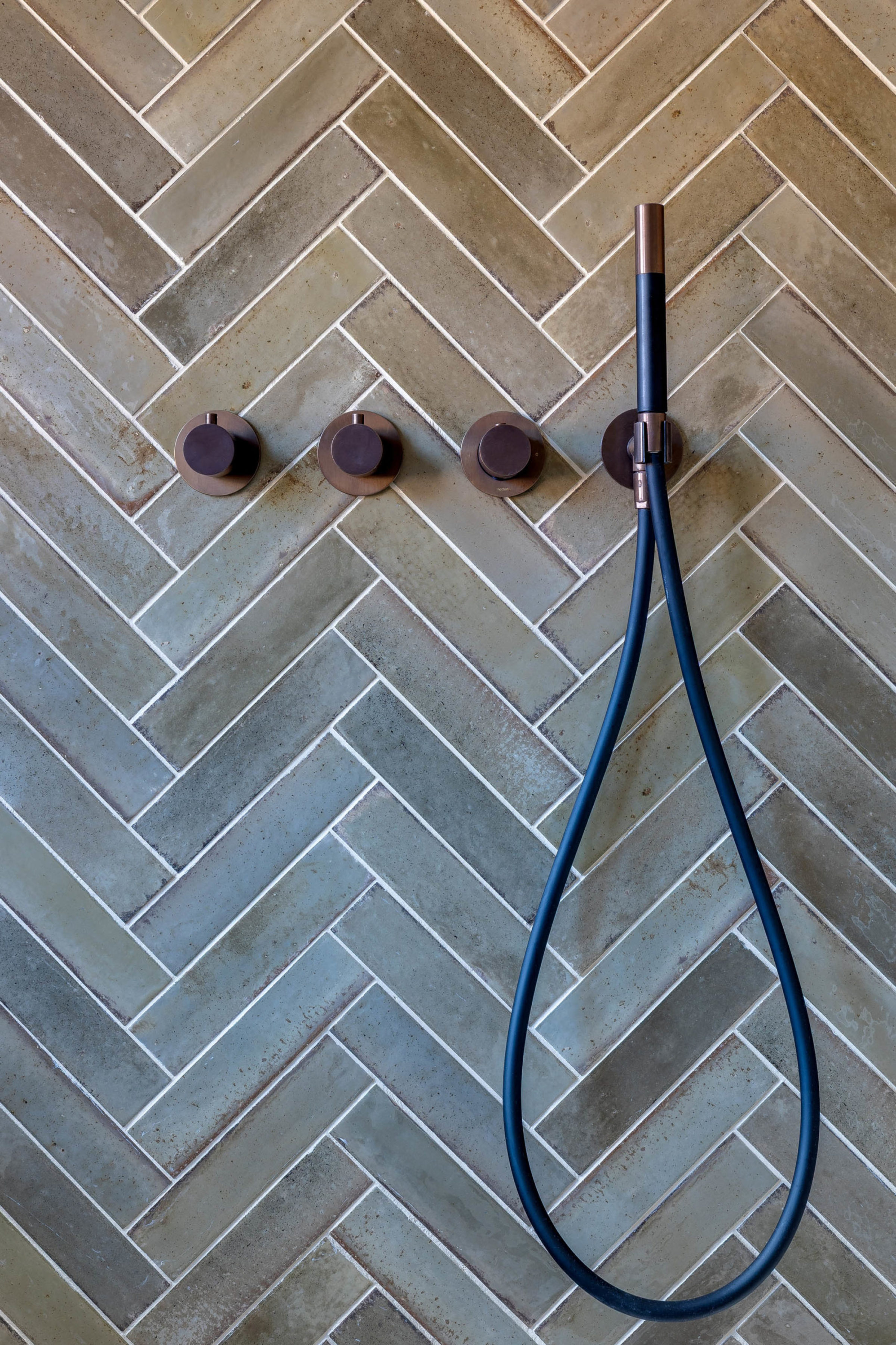Three round bronze control knobs in a horizontal row and a handheld shower with a black hose mounted on a green‑beige herringbone tiled wall.