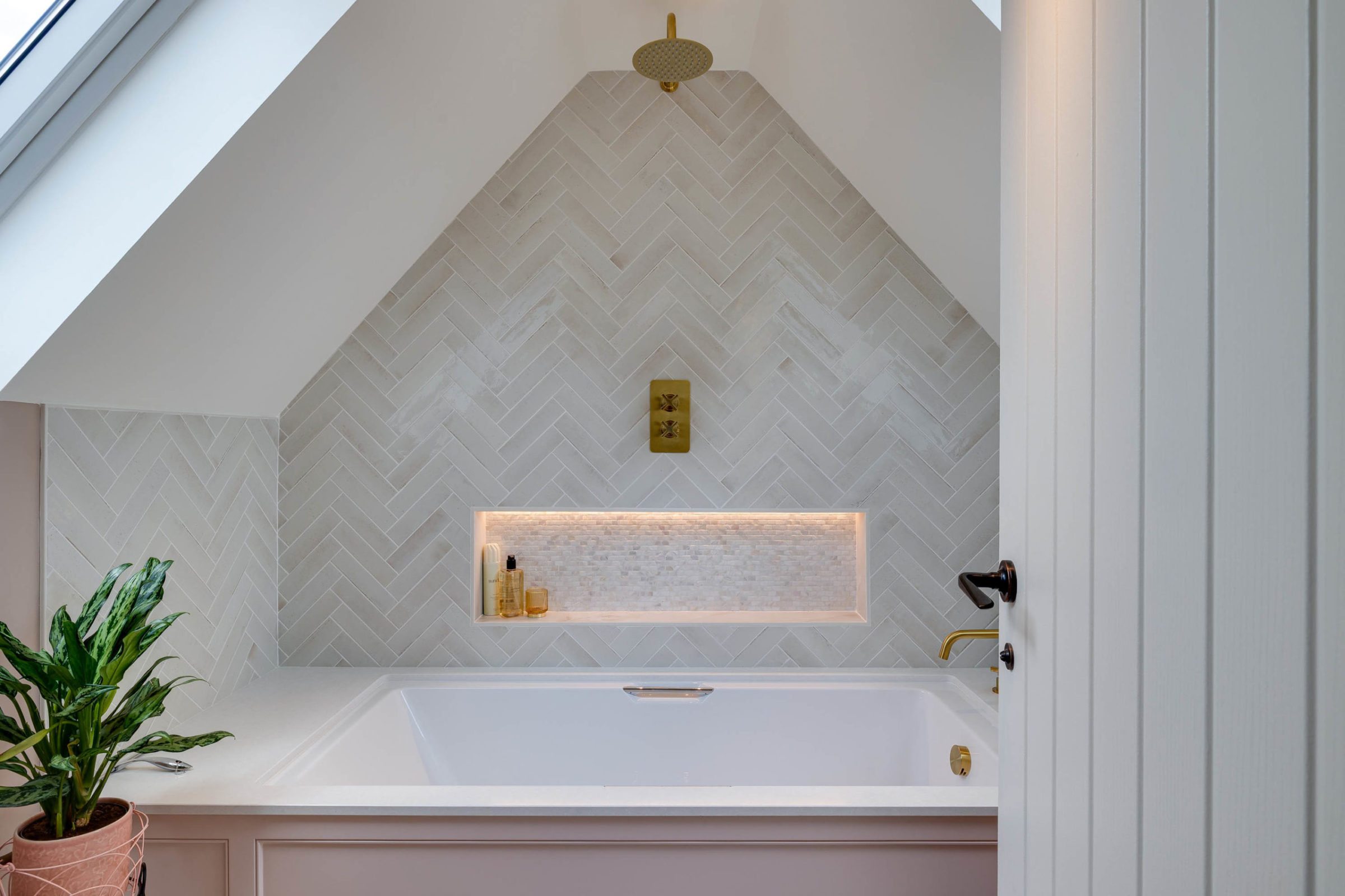 Recessed bathtub beneath a sloped ceiling clad in pale herringbone tiles, featuring a brass overhead shower and controls, an illuminated recessed niche with bottles, and a potted plant at the left corner.