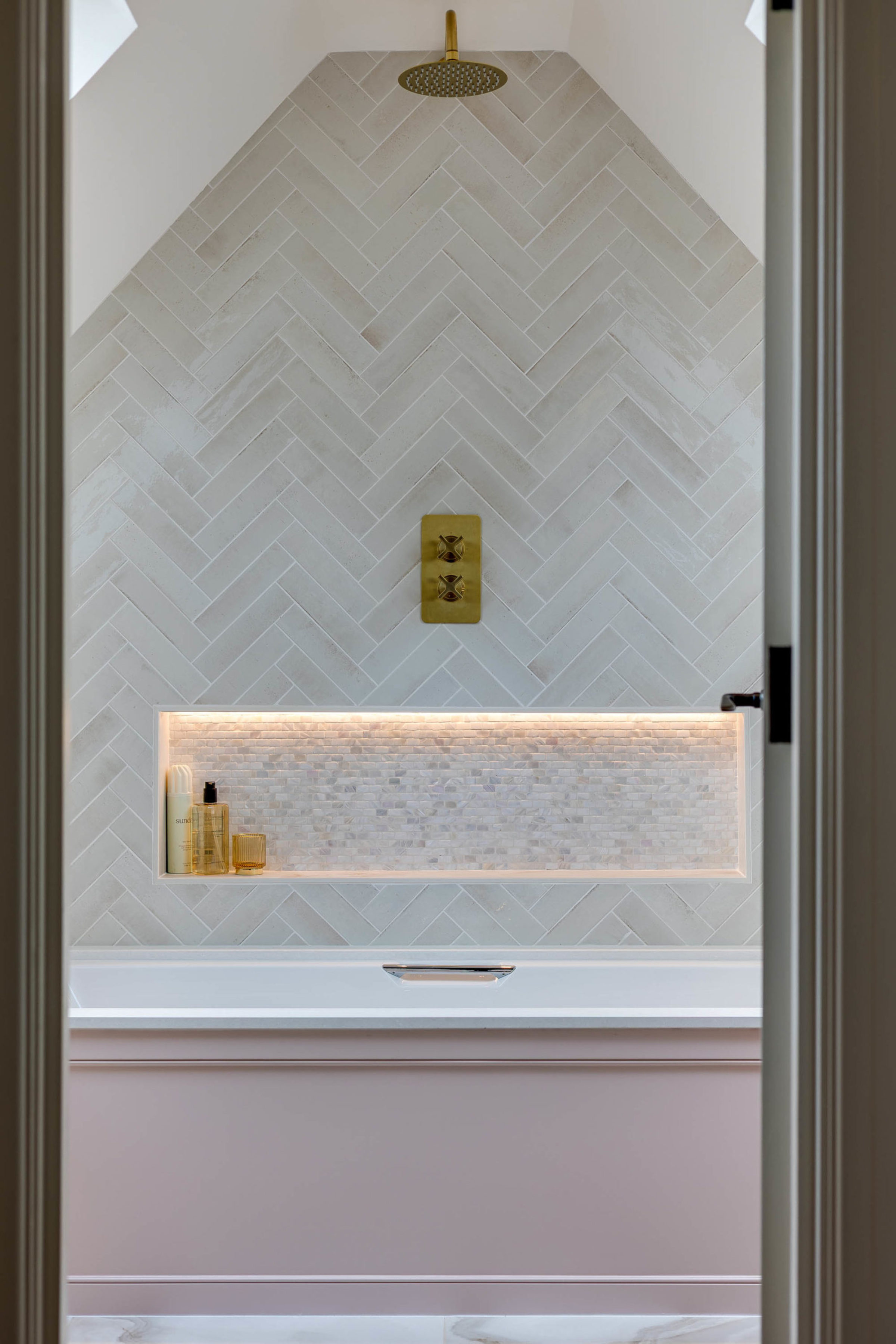 Light herringbone-tiled shower wall with a brass rain shower and matching brass controls above a lit recessed niche holding toiletries, with the pale pink bathtub edge visible below.