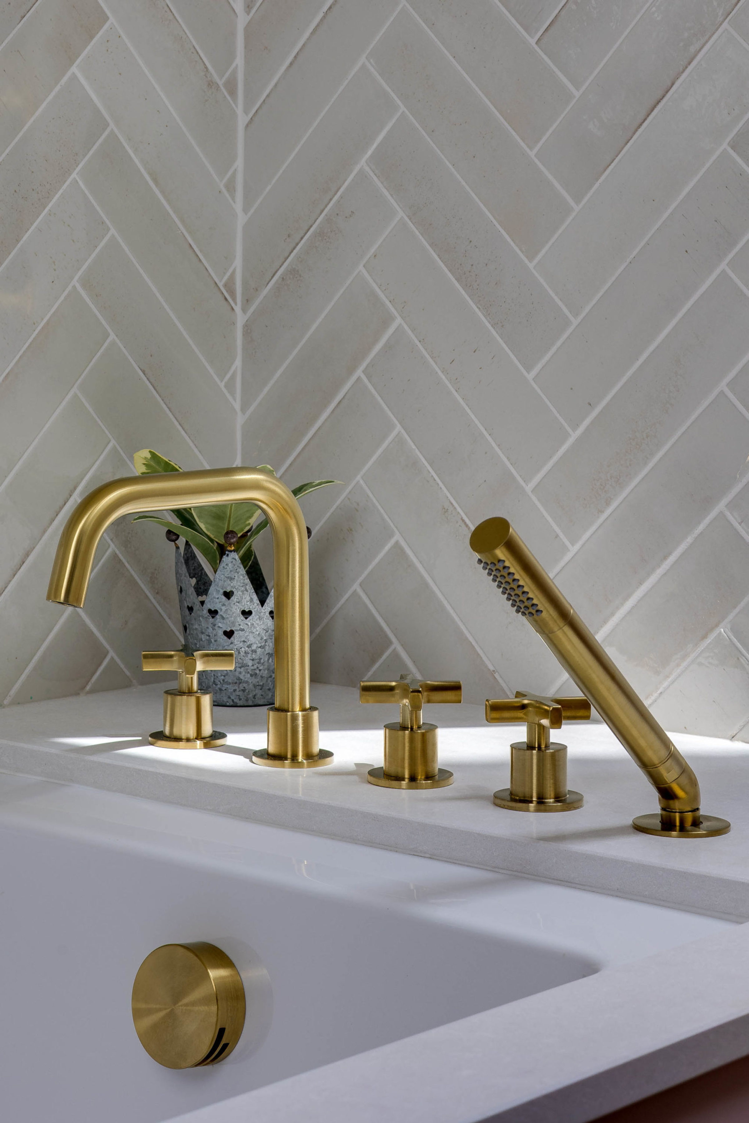 Brushed brass bathtub faucet, cross-handle controls and angled handheld sprayer installed on a white ledge with a small metal plant pot behind, set against pale grey herringbone tiles.