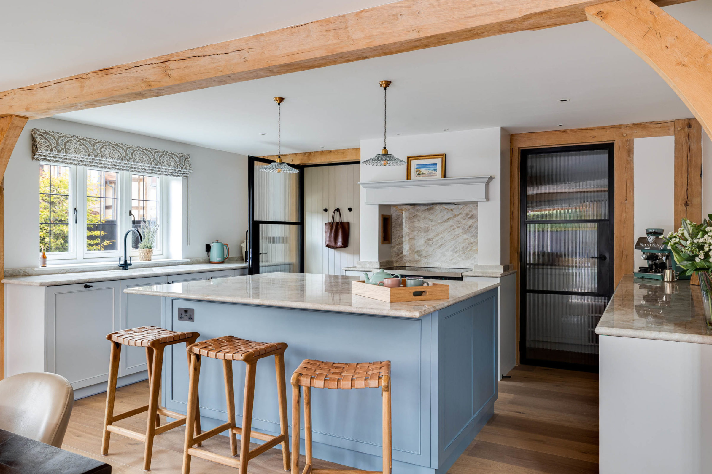 Bright farmhouse-style kitchen with exposed oak beams, a large blue central island topped with a marble countertop and wooden tray, three woven-leather bar stools, pendant lights overhead and a windowed sink area to the left.
