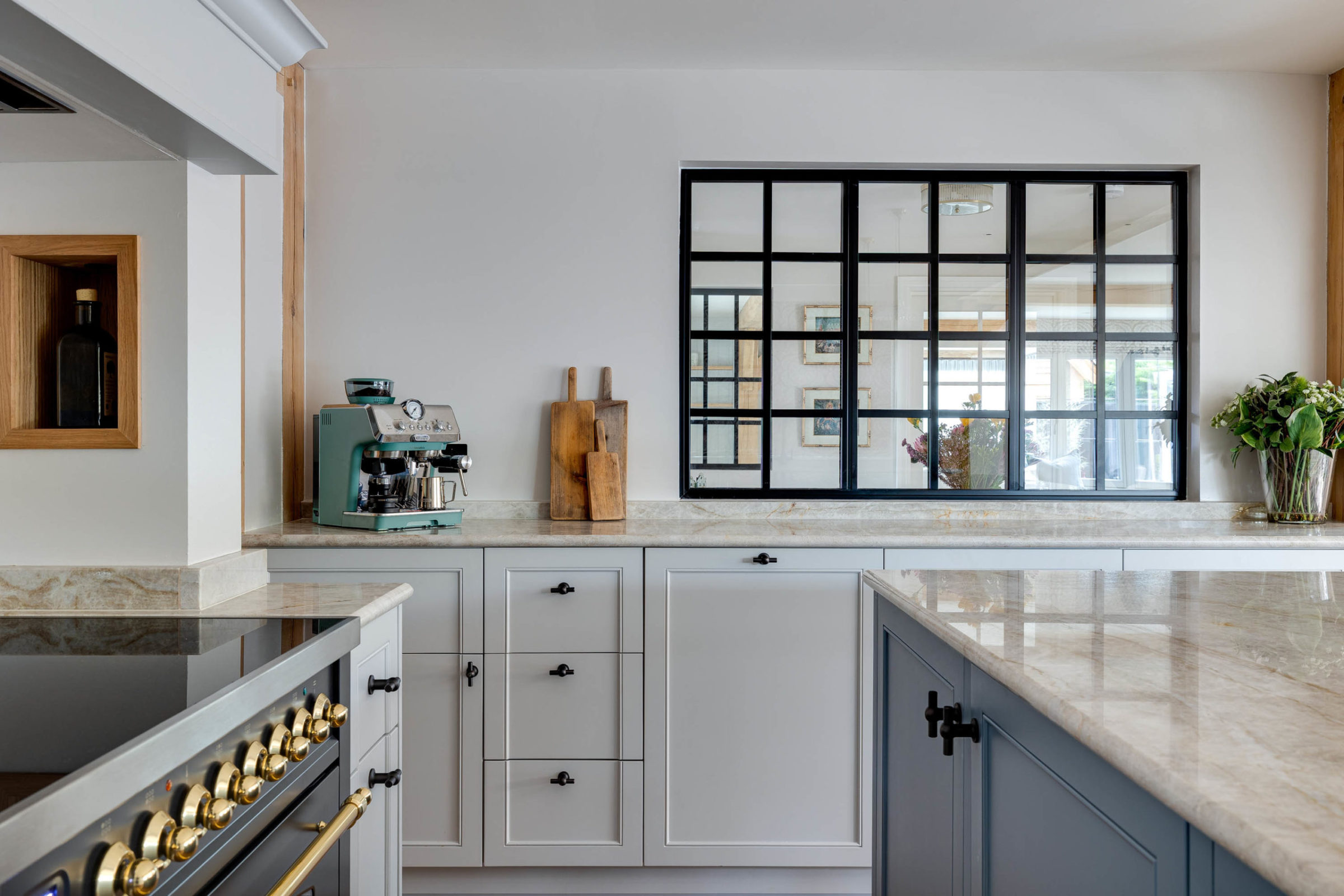 Bright modern kitchen with marble countertops and grey cabinetry, featuring a teal espresso machine and wooden cutting boards on the left, a large black-framed interior window reflecting the adjacent room, brass stove knobs in the foreground and a glass vase of flowers on the right.