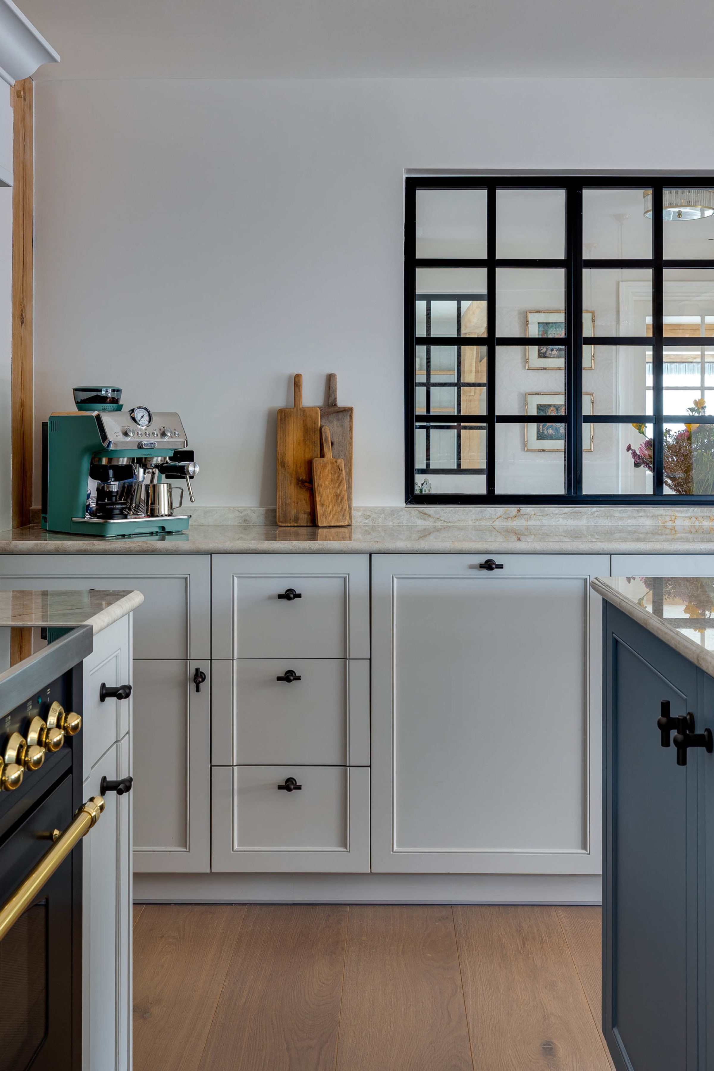 Marble kitchen countertop with a teal espresso machine on the left, three wooden cutting boards leaning against a pale wall, and a black-framed grid window revealing framed pictures and flowers in the adjoining room above white cabinetry.