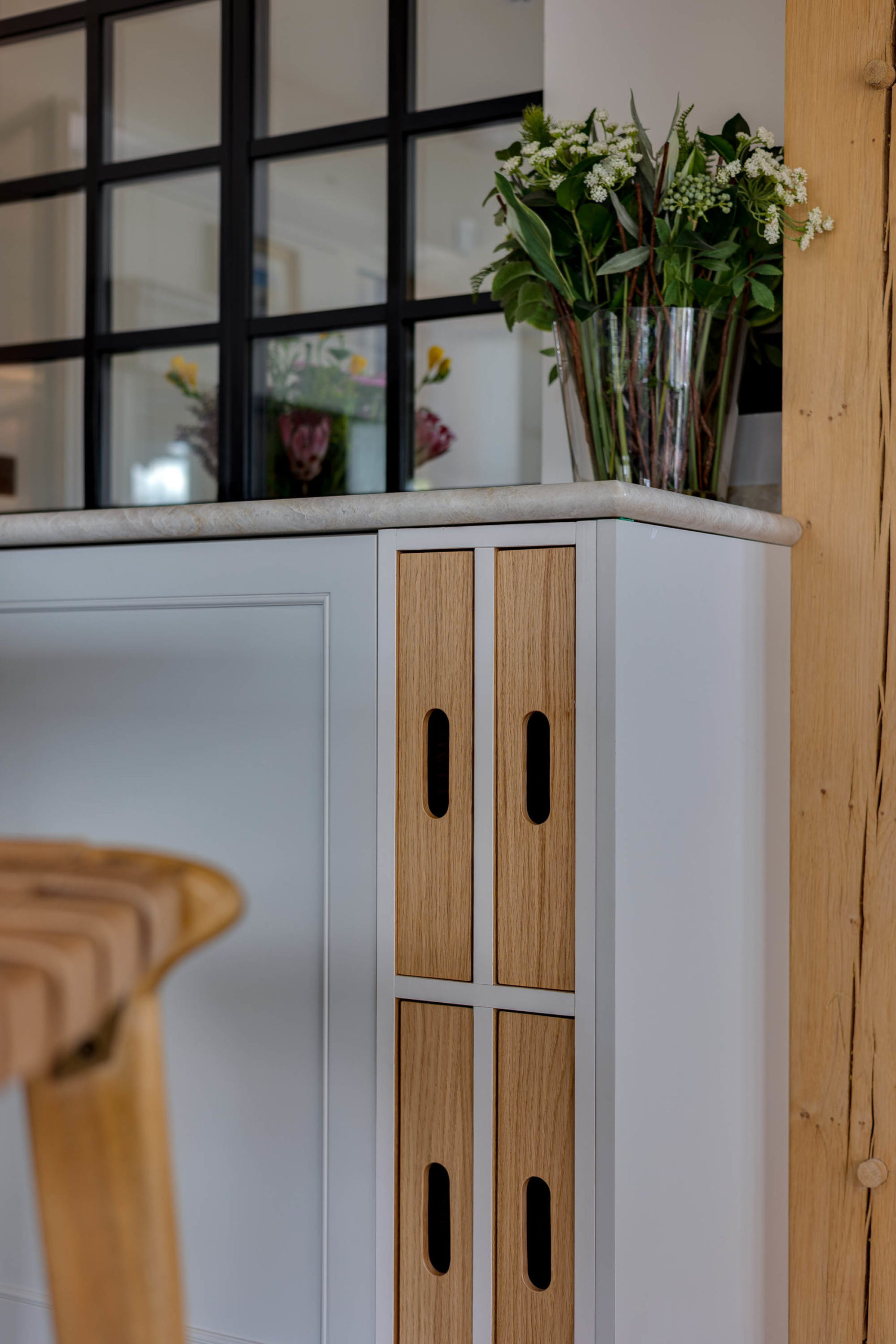 Corner of a light-grey kitchen island with a marble-effect countertop, vertical oak slats with oval cut-outs, a clear vase of green foliage and small white flowers on top, and a black-framed glass partition with a timber beam in the background.