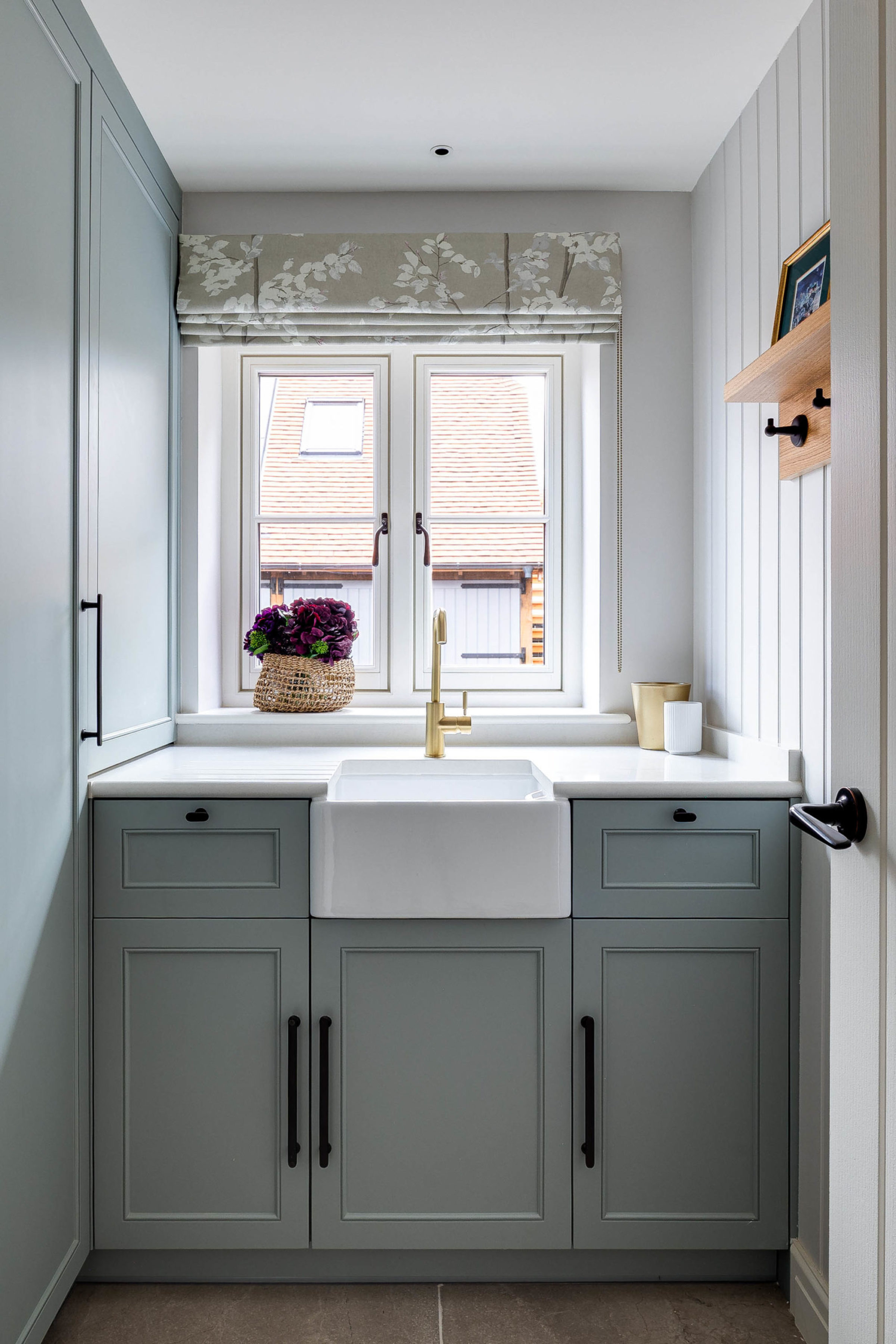 Small laundry-style sink area with a white apron-front farmhouse sink and brass tap beneath a double window dressed with a folded floral Roman blind, flanked by pale blue-grey shaker cabinets and a windowsill basket of purple flowers, with a wooden shelf and hooks on the paneled right wall.