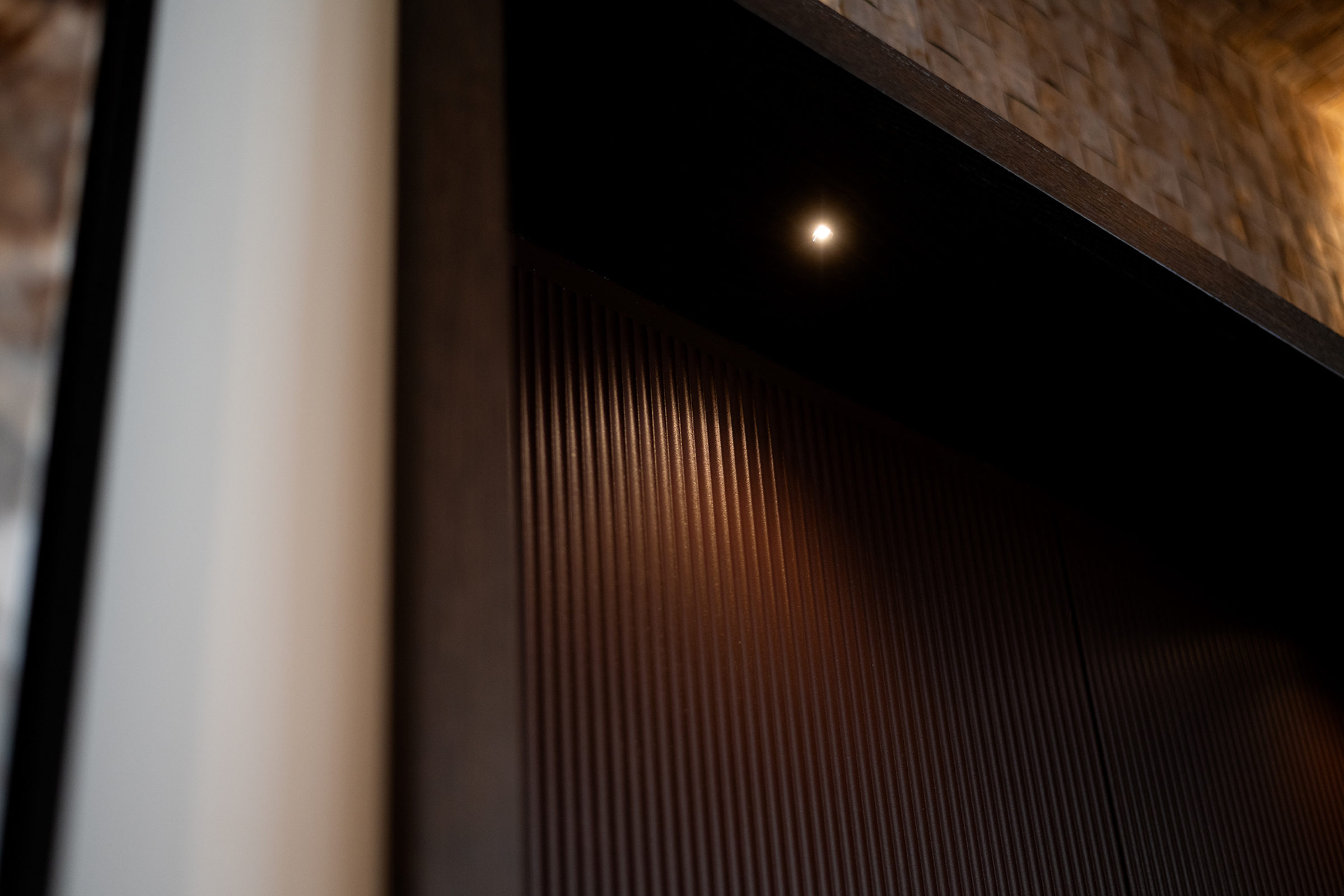 Close-up of dark vertical-ribbed wood paneling under an angled shelf with a small recessed spotlight casting a warm glow and a blurred pale column at the left edge.