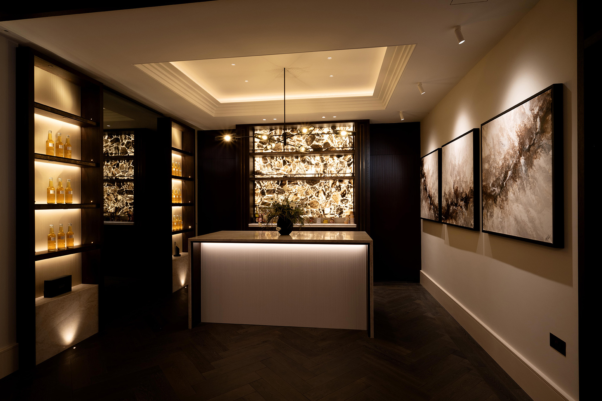 Warm, dimly lit modern reception or bar interior with a central white-fronted island under recessed ceiling lighting and a slim black pendant, a backlit onyx feature wall behind the counter, illuminated shelving of amber bottles to the left, and three large abstract framed paintings along the right wall.