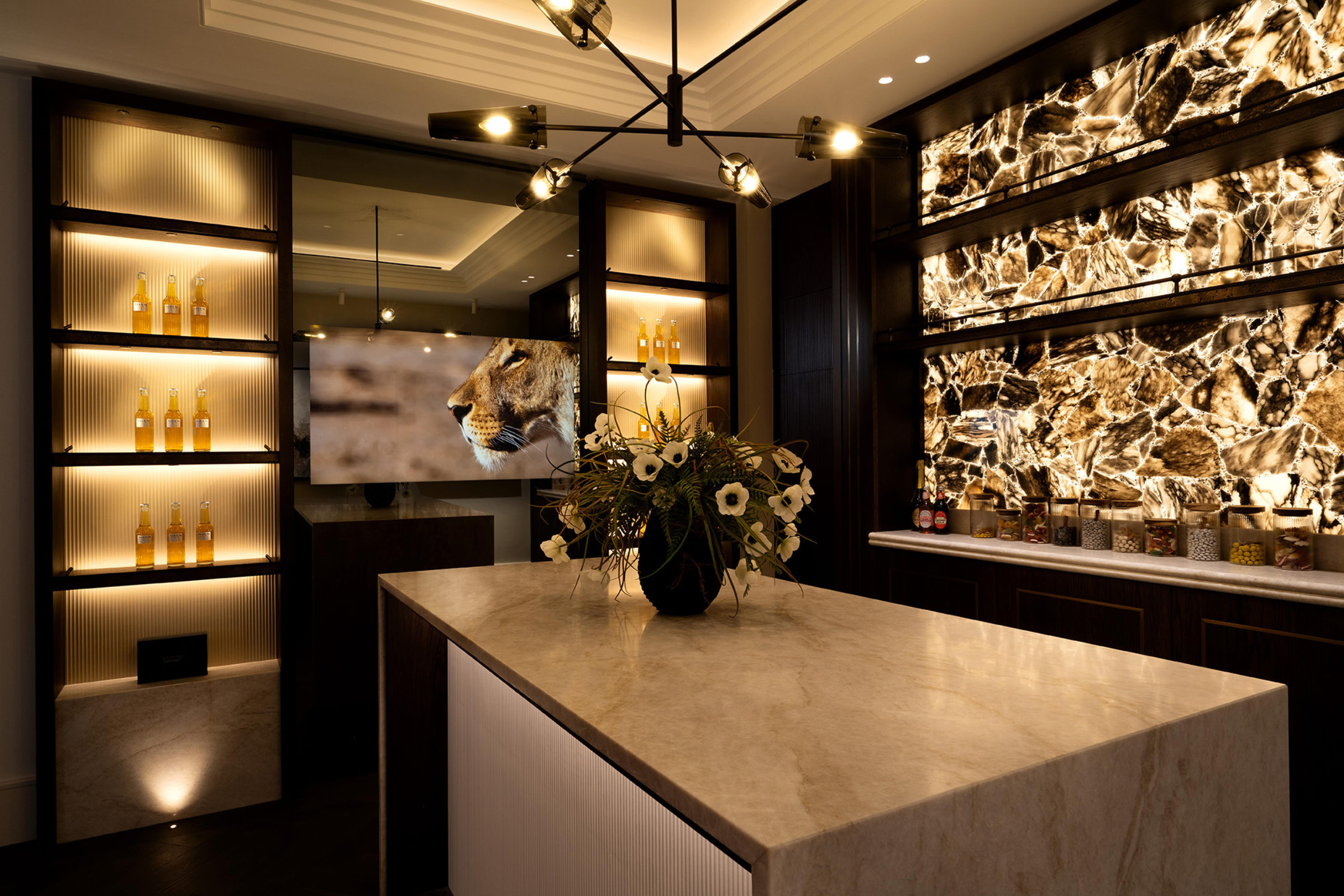 Luxurious dimly lit bar interior with a marble-topped island and vase of white flowers, backlit stone feature wall and illuminated shelves of bottles, a modern multi-arm chandelier overhead and a TV screen showing a lion's profile.