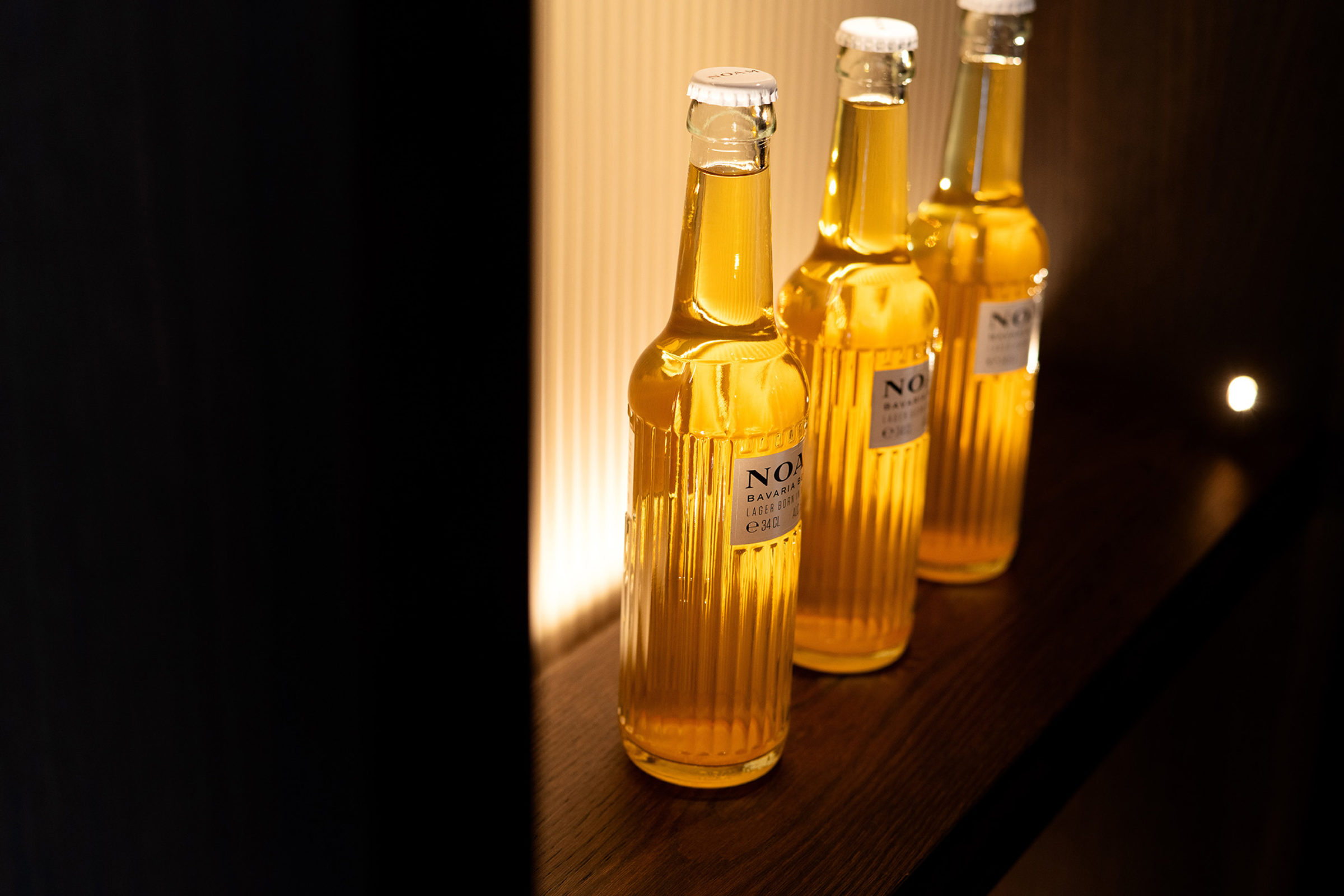 Three tall ribbed glass bottles of golden lager with white caps standing on a wooden shelf, warmly backlit by vertical lighting against a dark background.