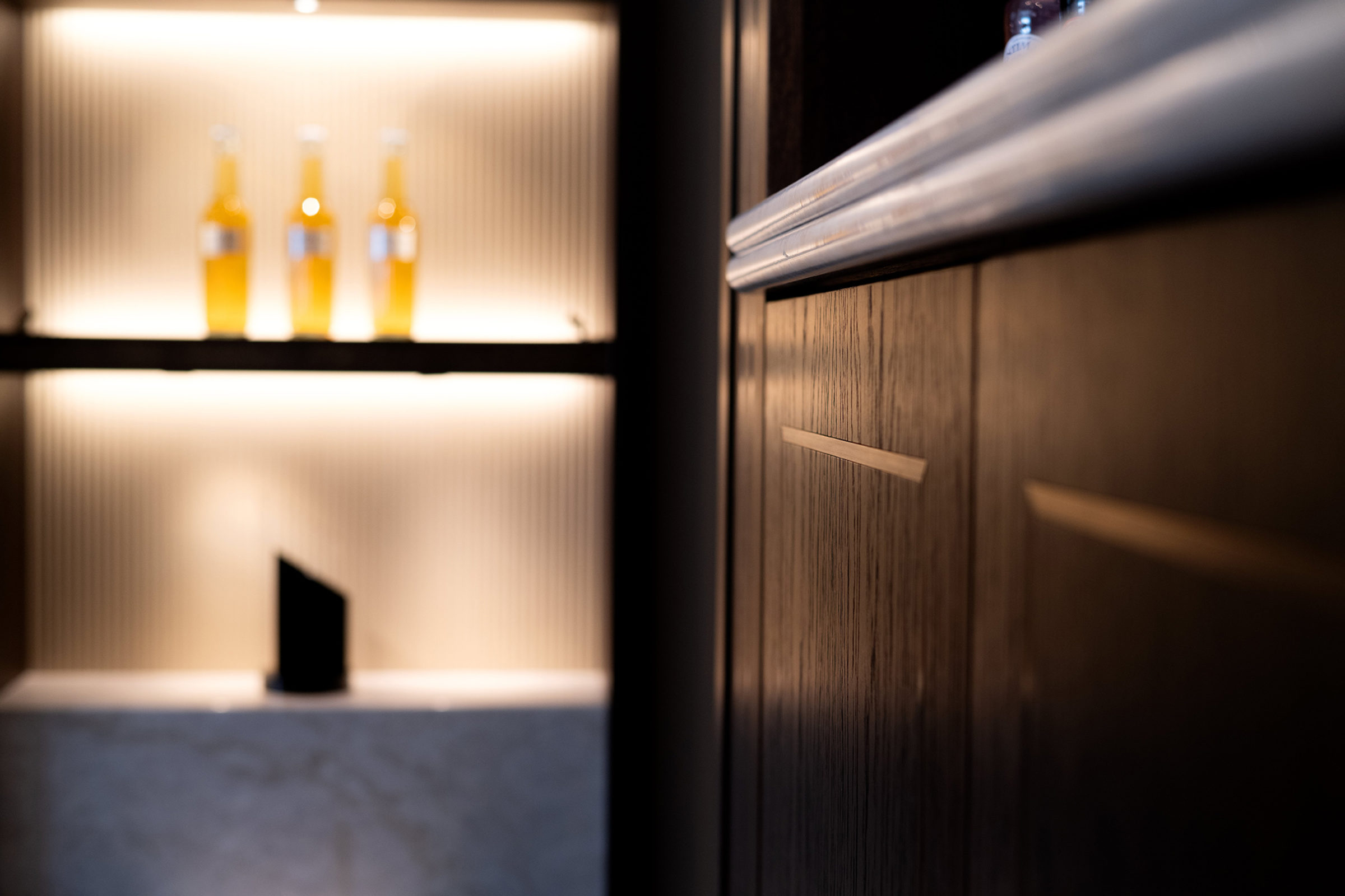 Close-up of a wooden cabinet edge and metal countertop trim in sharp focus with a softly lit shelf of three blurred amber bottles and ribbed back panel in the background.