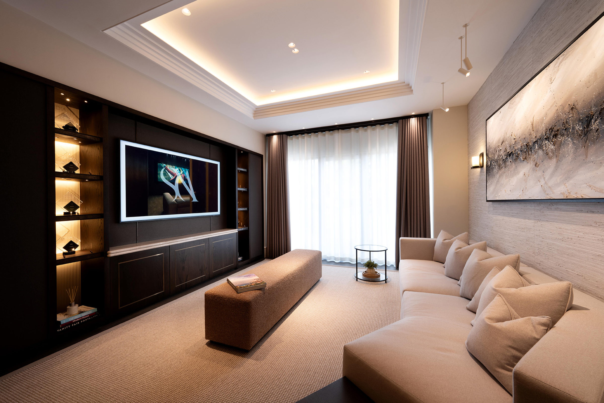 Contemporary living room with a large beige sectional sofa and cushions, a central upholstered bench with a book, a wall-mounted TV set into dark cabinetry with backlit display shelves, floor-to-ceiling curtains, a round glass side table with a plant, and a large abstract painting on a textured wall beneath warm recessed tray ceiling lighting.